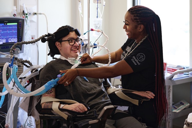 A nurse making adjustments to a patients ventilator equipment. Both are making eye contact and smiling at each other.