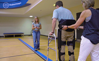 Two therapists helping a male client work on walking with a walkher and probiotic exoskeleton attached to his waist.