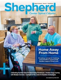 Three people in a kitchen: a person in a wheelchair with a blanket, a standing woman in a sweater, and a seated man in a t-shirt, smiling at each other.