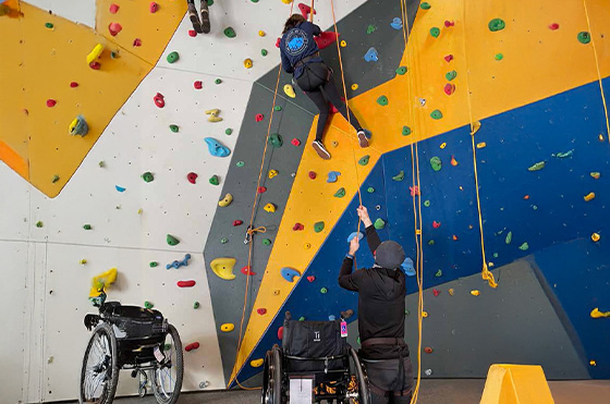 A person in a wheelchair climbs an indoor rock wall with support from another individual. Nearby, another wheelchair and climbing equipment are visible. The wall is colorful with various holds and routes.