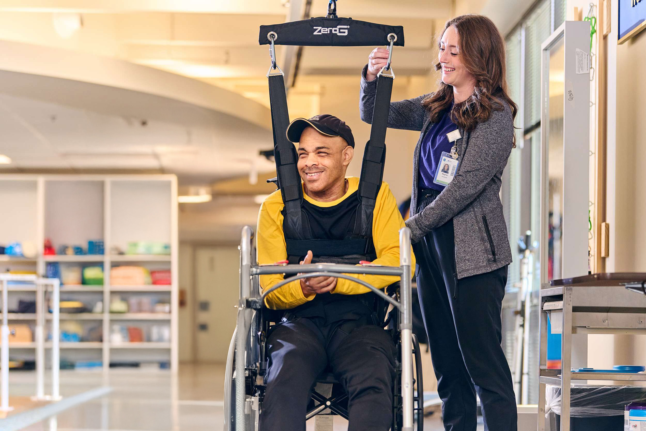 Two women assist a man seated in a wheelchair with a harness system. They are in a bright indoor setting, smiling and engaged in supportive interaction. The equipment and setting suggest a therapeutic or rehabilitation environment.