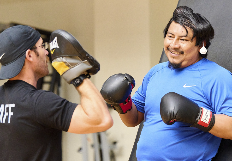 A man in a blue shirt with black boxing gloves smiles while facing a second man in a black 