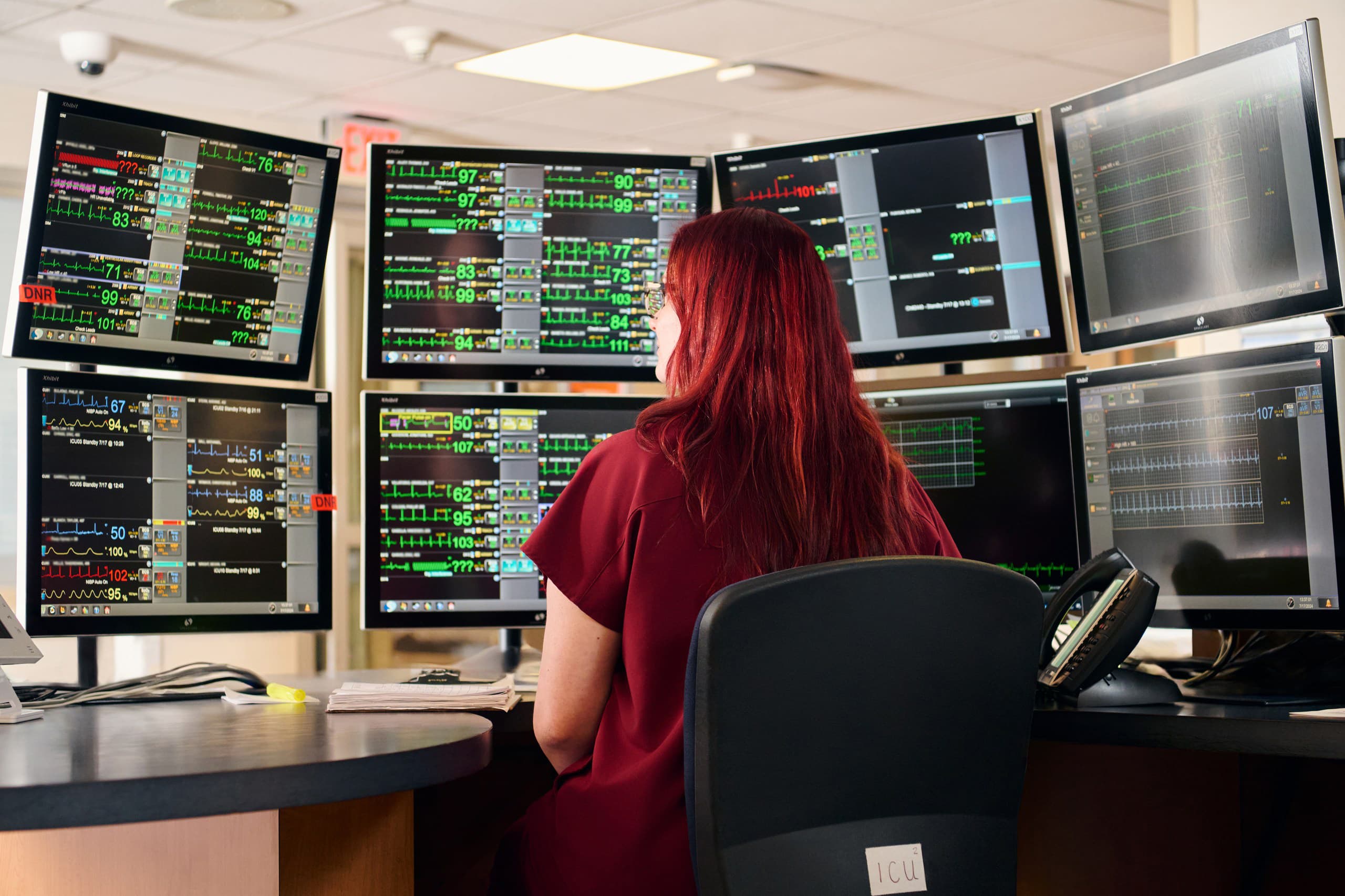 A person with red hair sits in a control room, facing multiple computer screens displaying various data and charts. They are seated in a modern office chair, and a landline phone is visible on the desk.