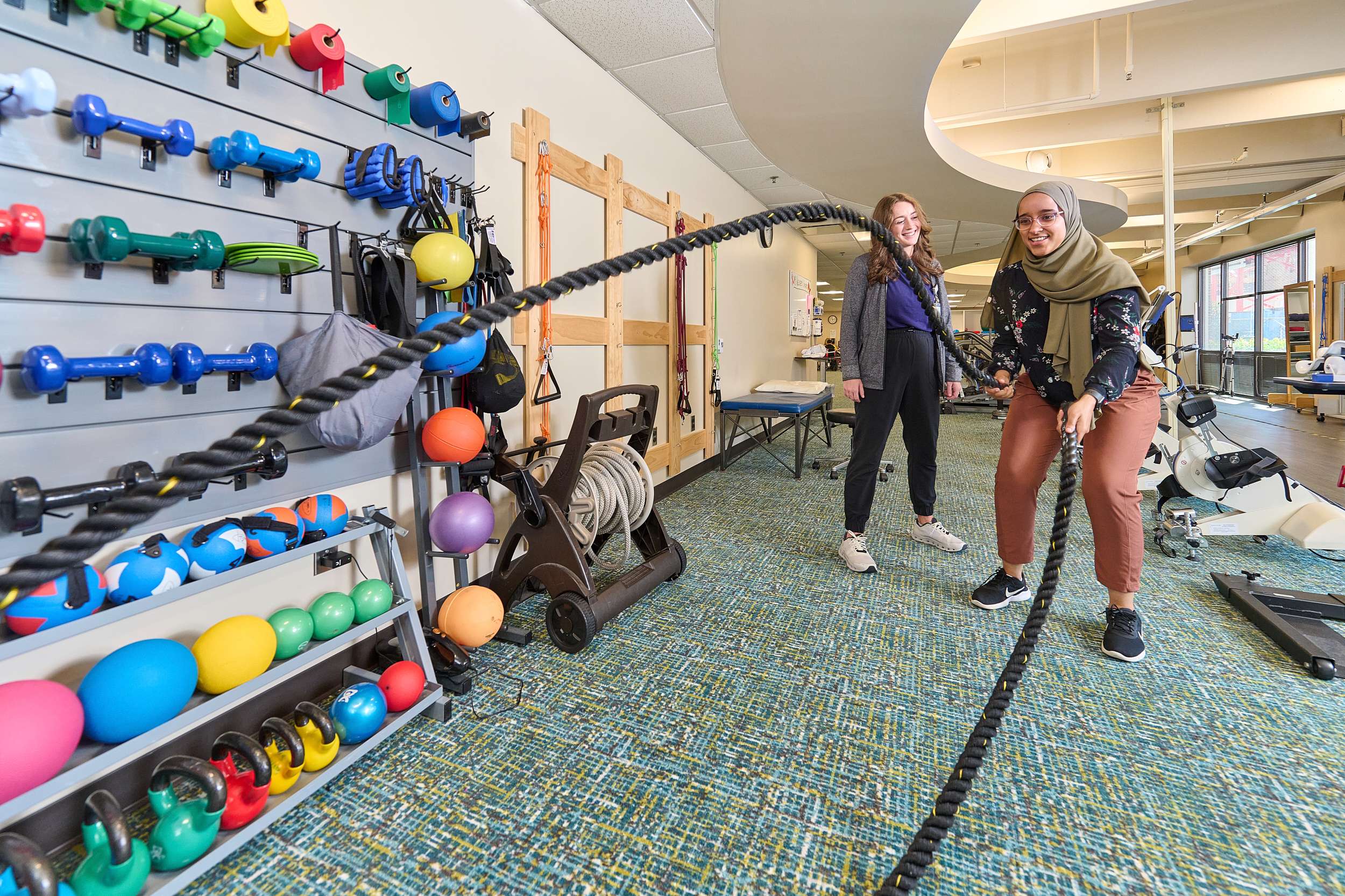Two women are exercising in a gym. One is using battle ropes while the other watches and smiles. The room has a wall rack with colorful dumbbells, kettlebells, and medicine balls, and various fitness equipment in the background.