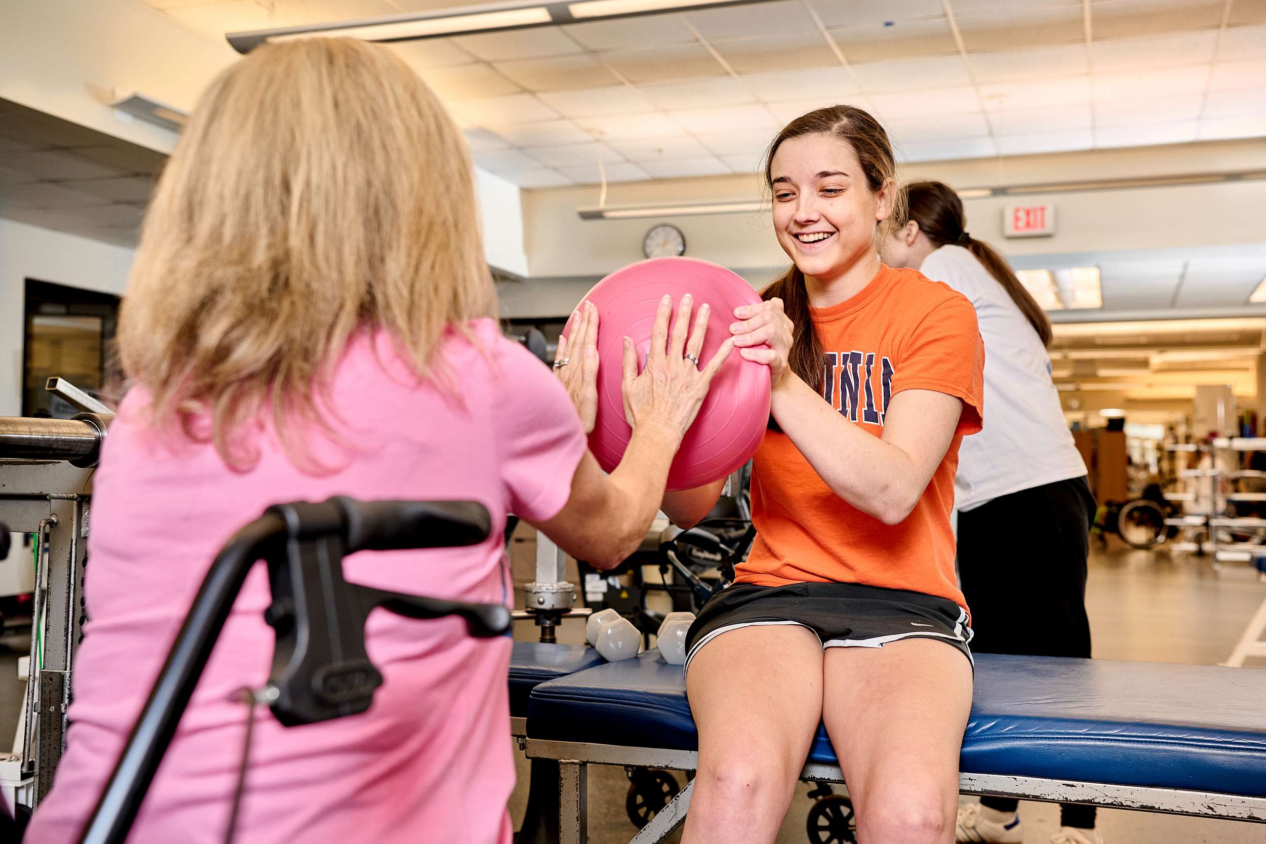 A woman in a pink shirt assists a smiling young woman in an orange shirt as they hold a pink exercise ball between them in a gym. The young woman is sitting on a therapy bench. Equipment and another person are visible in the background.