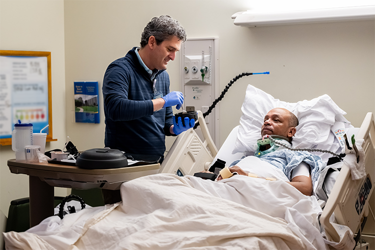 A man lies in a hospital bed on a ventilator, while a volunteer stands beside the bed, feeding him.