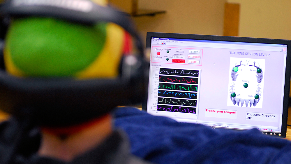 A research participant with a magnetic sensor attached to their tongue is testing tongue-controlled technology. In front of them, a laptop screen displays charts and a diagram of a mouth. A red prompt on the screen reads: Freeze your tongue! You have 5 seconds left.