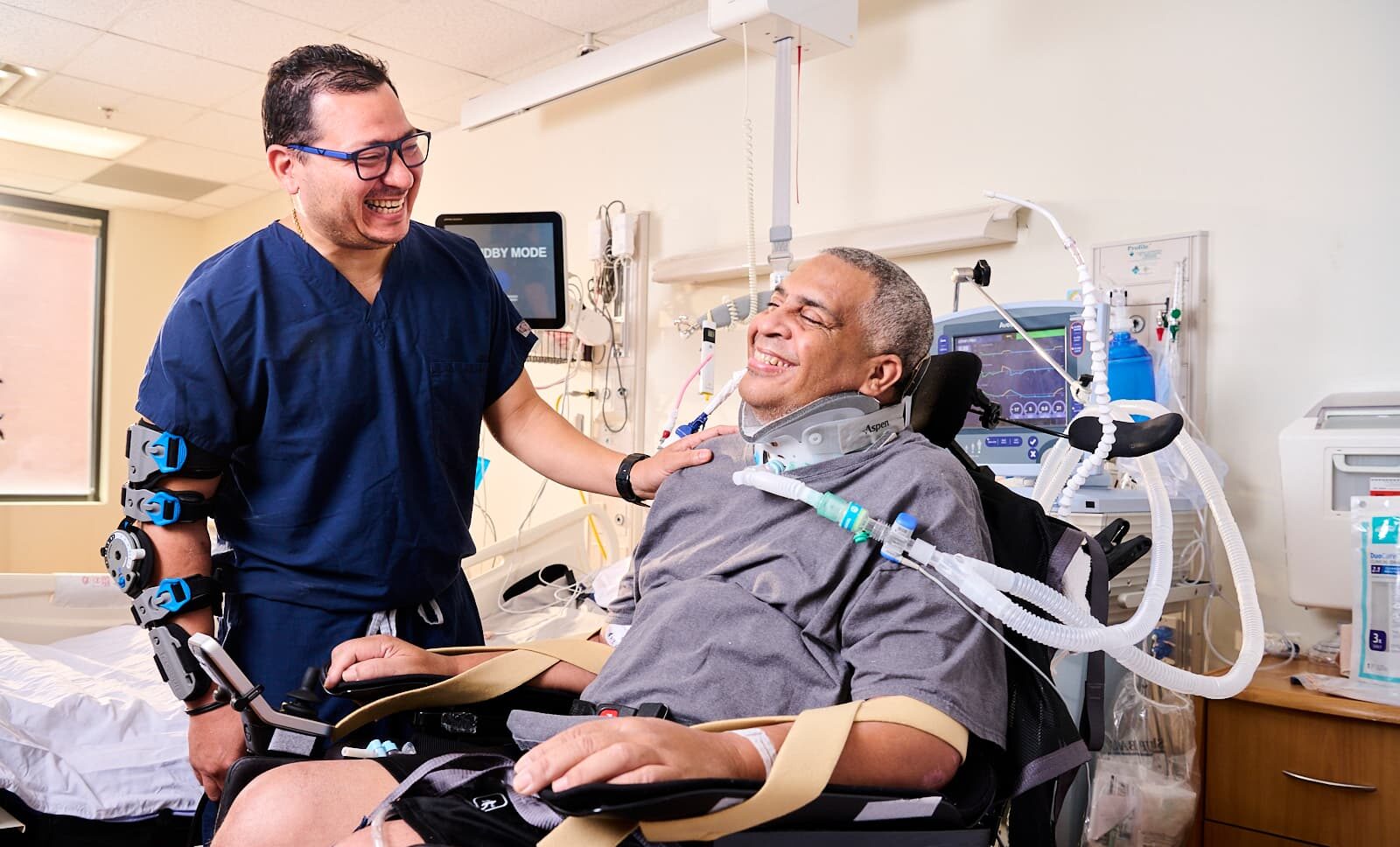 A nurse in navy scrubs stands smiling beside a seated hospital patient in a supportive chair and neck brace, connected to medical equipment. The scene takes place in a brightly lit hospital room, creating a warm and caring atmosphere.