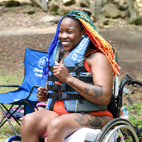 Female with colorful hair smiles big while seated in her wheelchair wearing a life jacket.