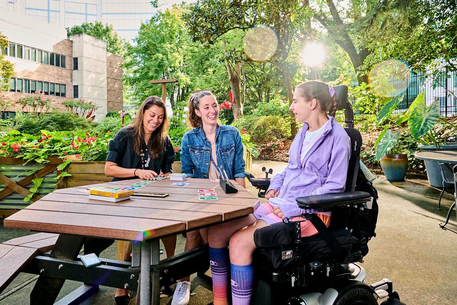 Three women are sitting at an outdoor picnic table. Two of them are playing a card game, while the woman on the right, using a motorized wheelchair, is engaged in the conversation. Surrounded by lush greenery with sunlight filtering through the trees, they enjoy their team workspace in nature.