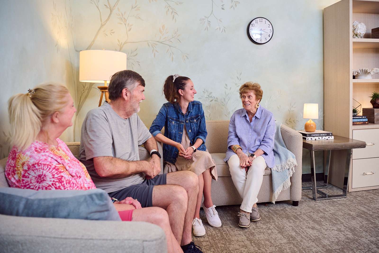 In a well-lit waiting room with neutral decor, four people, which includes a patient and their family, sit engaged in a discussion.