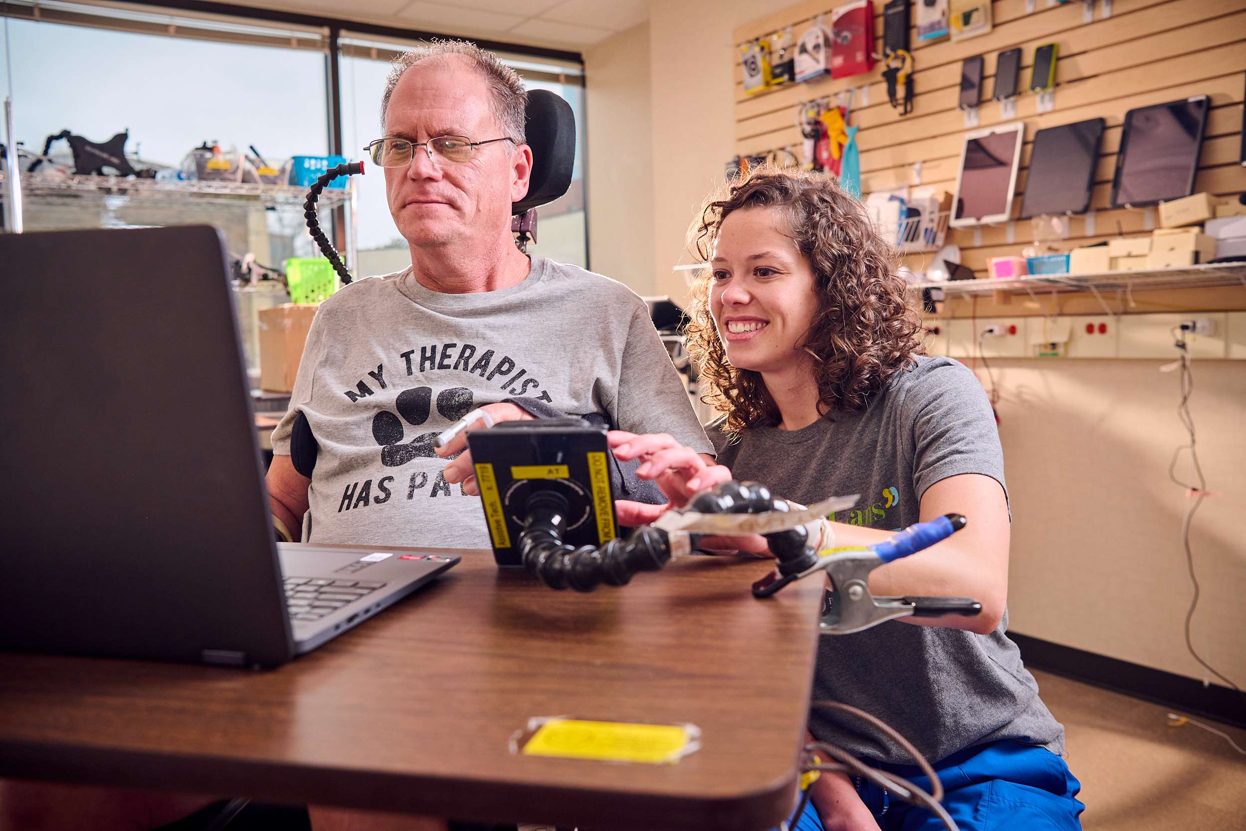 A man in a wheelchair uses a computer with an adaptive device, assisted by a smiling woman. They are in a room with technology equipment on the wall. The man's shirt humorously reads 