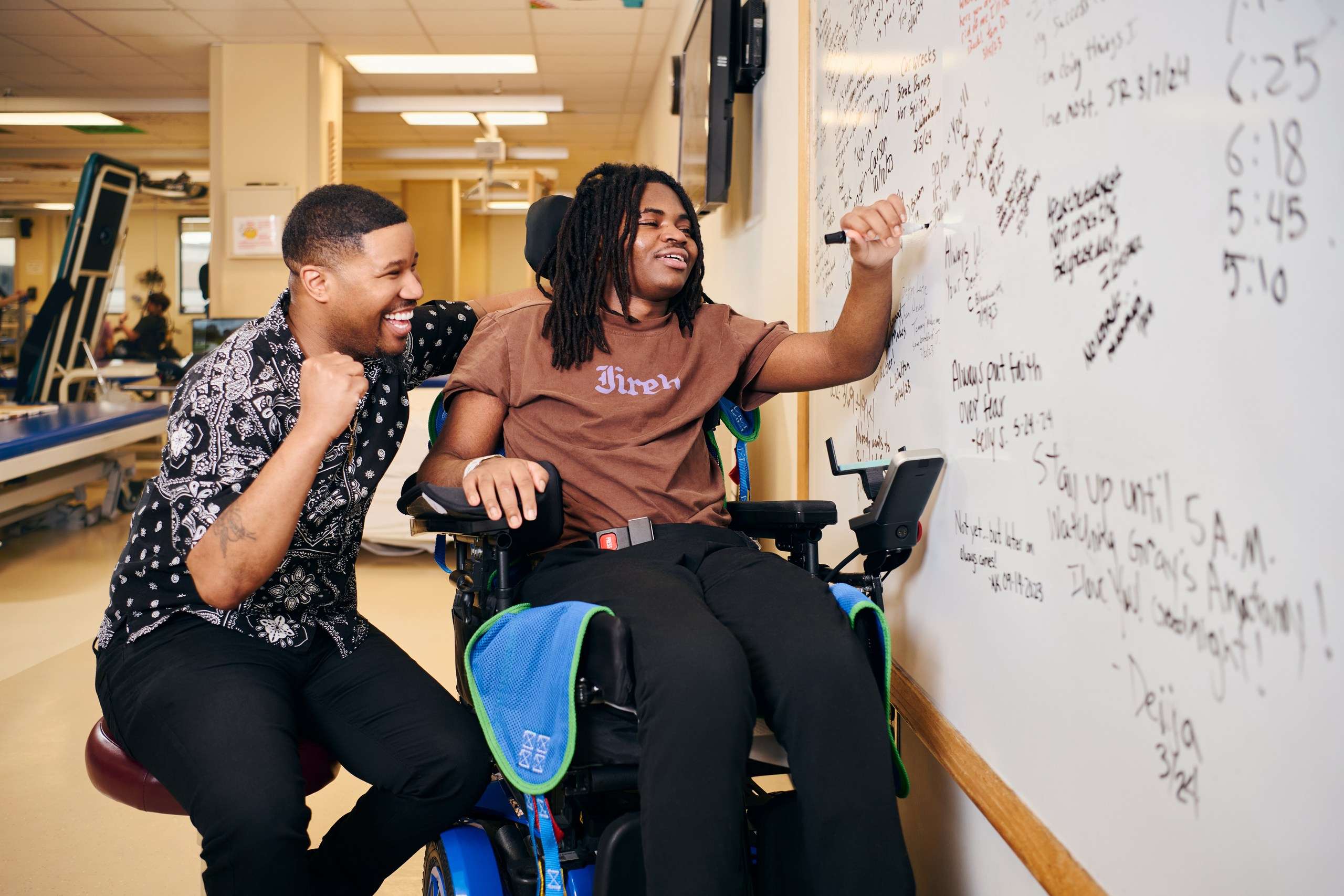 Cedric Maddox assists Jakoi Horne, seated in a motorized wheelchair, as he writes a note of encouragement on a large whiteboard filled with various notes and doodles from other SCI adolescent graduates.