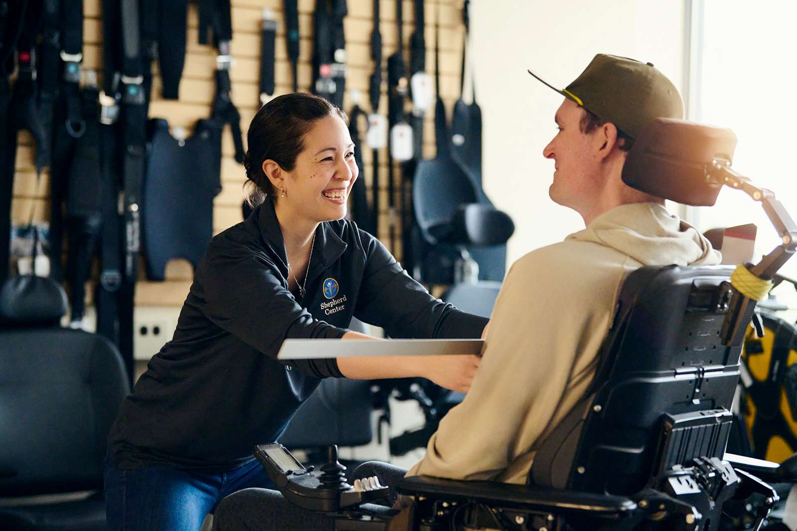 A woman in a dark jacket is smiling and adjusting the armrest of a man sitting in a motorized wheelchair. They are in a room adorned with various mobility aids on the wall. The atmosphere is friendly and supportive.