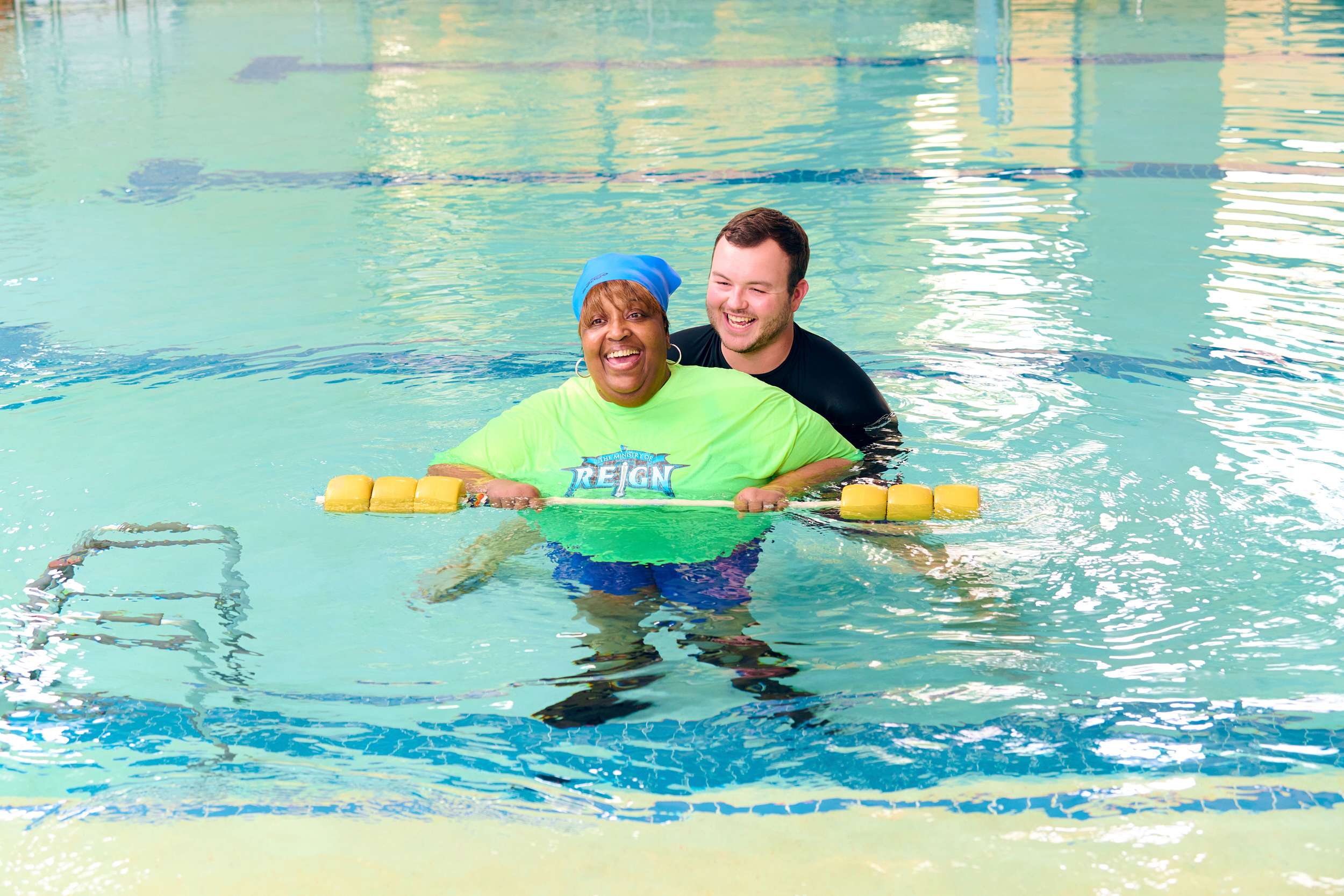 An older adult smiles in a pool, supported by a younger adult behind. They are holding a yellow flotation device during a water exercise session, with the pool lanes visible in the background.