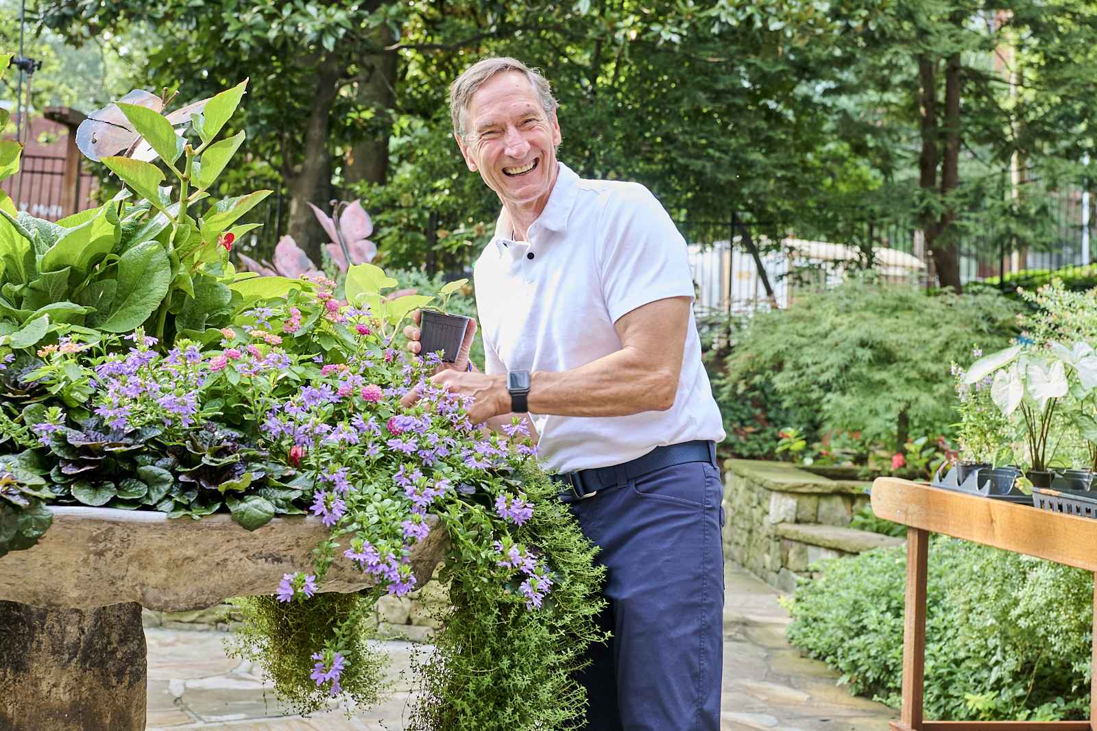 A person in a white polo shirt smiles while tending to a large stone planter filled with vibrant green plants and purple flowers. The background features a lush garden with various plants and trees on a sunny day.