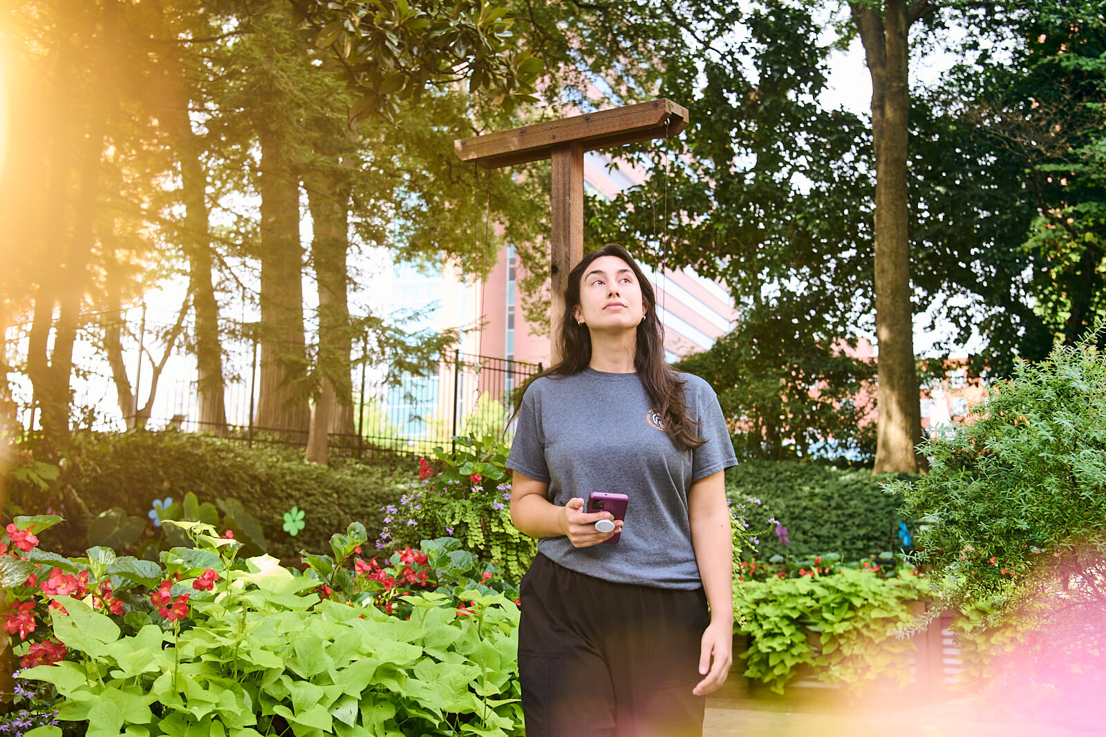 A woman walks in a garden, holding a coffee cup. She gazes upward, surrounded by lush greenery and colorful flowers. Sunlight filters through the trees, creating a warm, serene atmosphere.