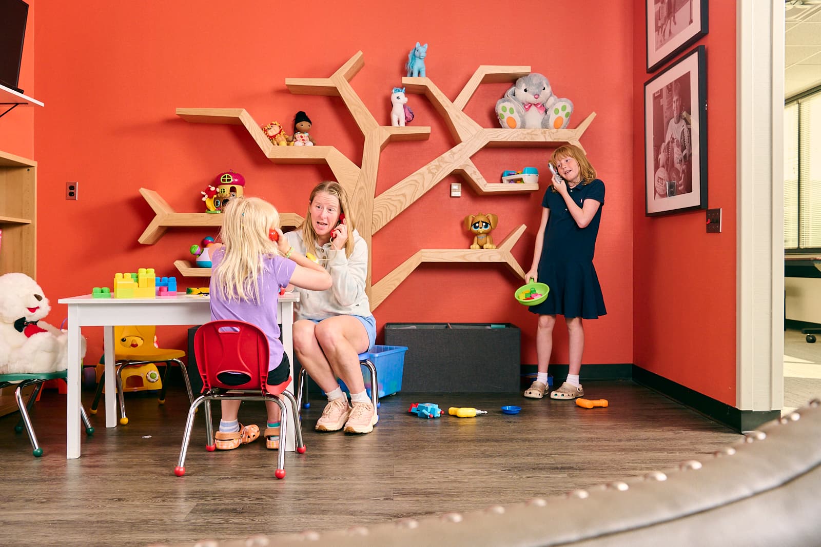 A woman and a child sit at a small table in a room with red walls and a tree-shaped shelf holding toys. Another woman stands nearby, observing. The floor is scattered with more toys, creating a playful and colorful environment.