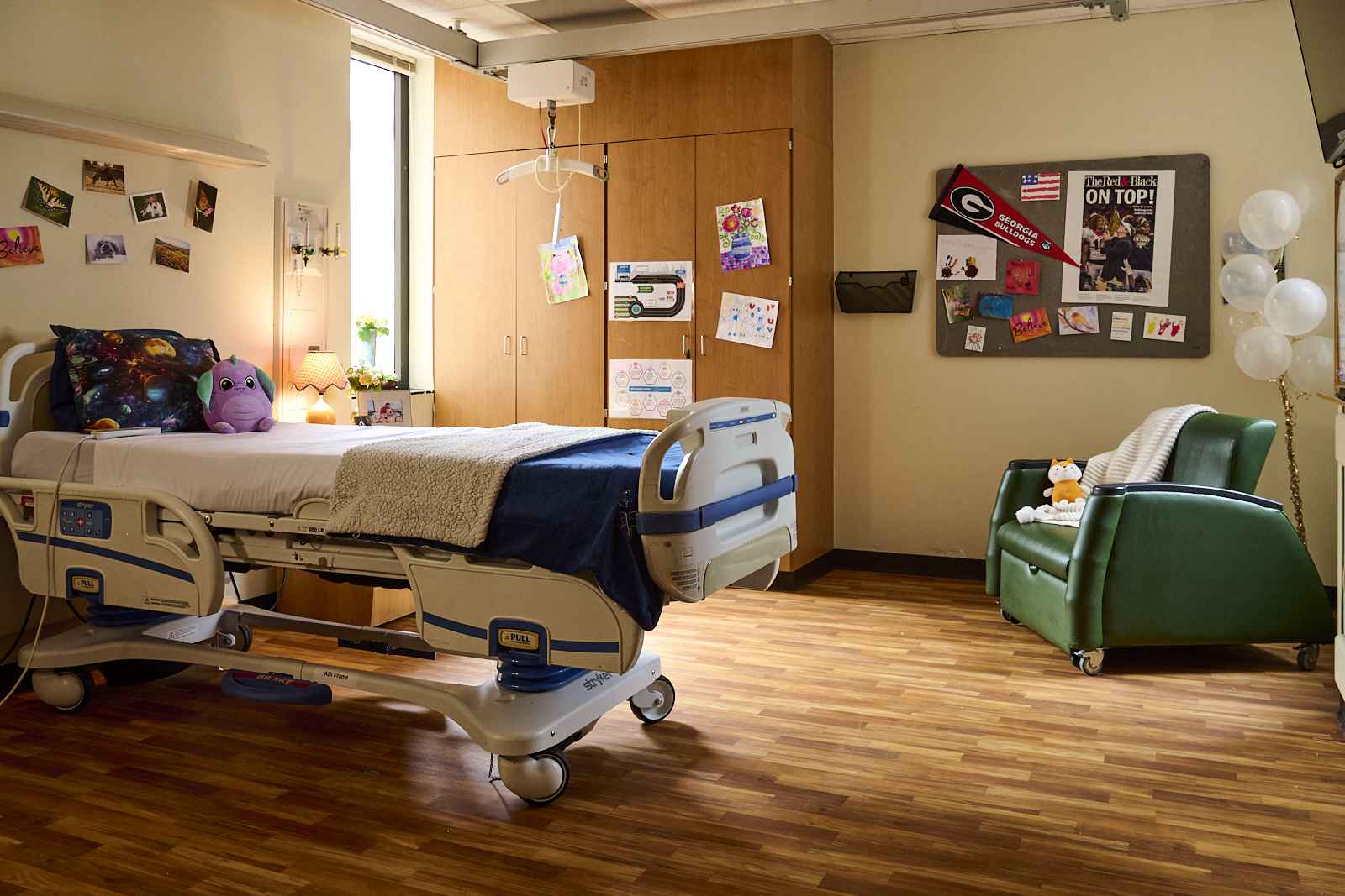 A cozy hospital room with wood flooring features a patient bed adorned with plush toys, colorful blankets, and a pillow. The walls are decorated with photos, drawings, and flags. Next to the bed is a green chair with a fox plushie and a bouquet of balloons.