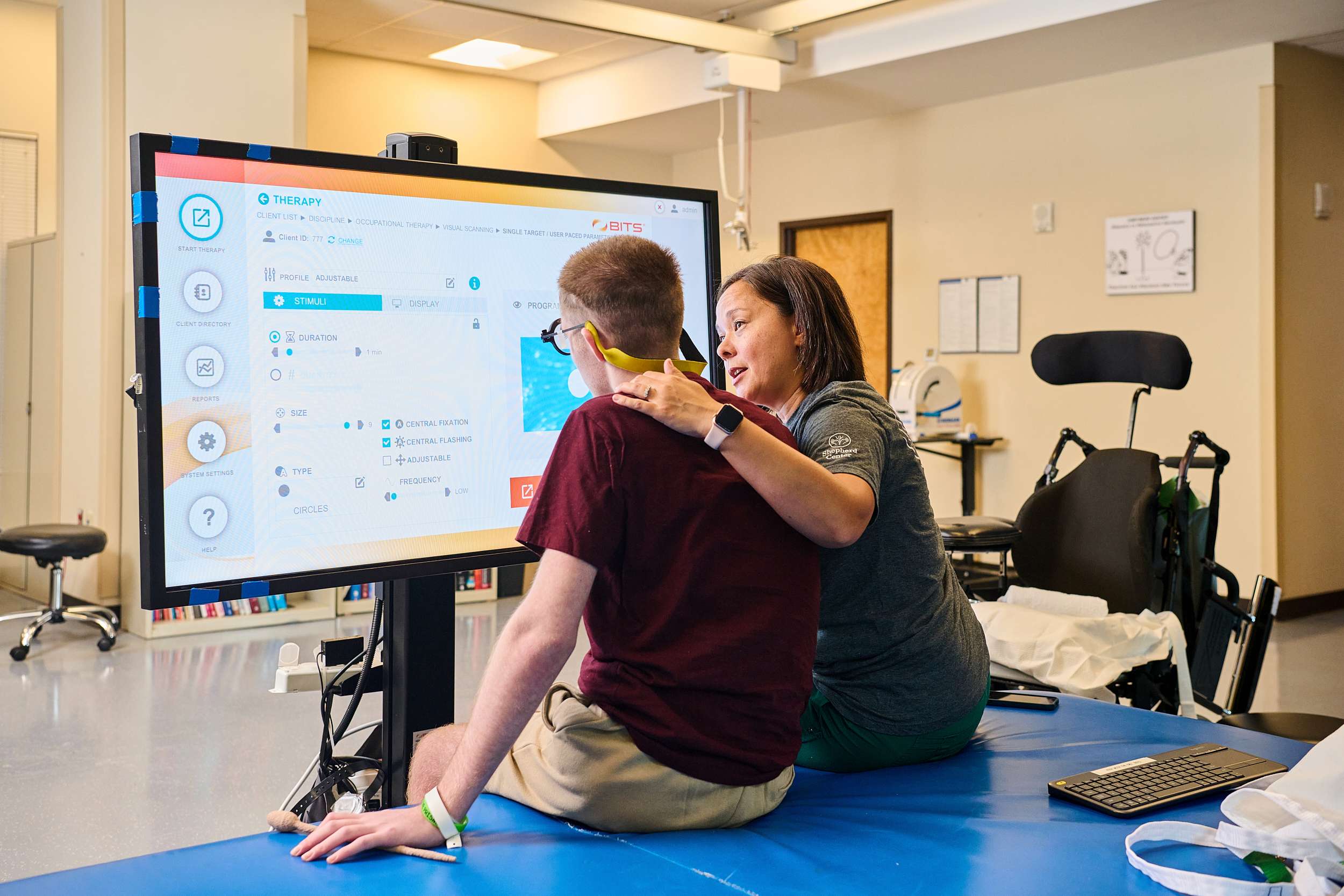 Occupational therapist puts her arm around the shoulder of an adolescent patient. In front of them is a large television screen used for therapy.