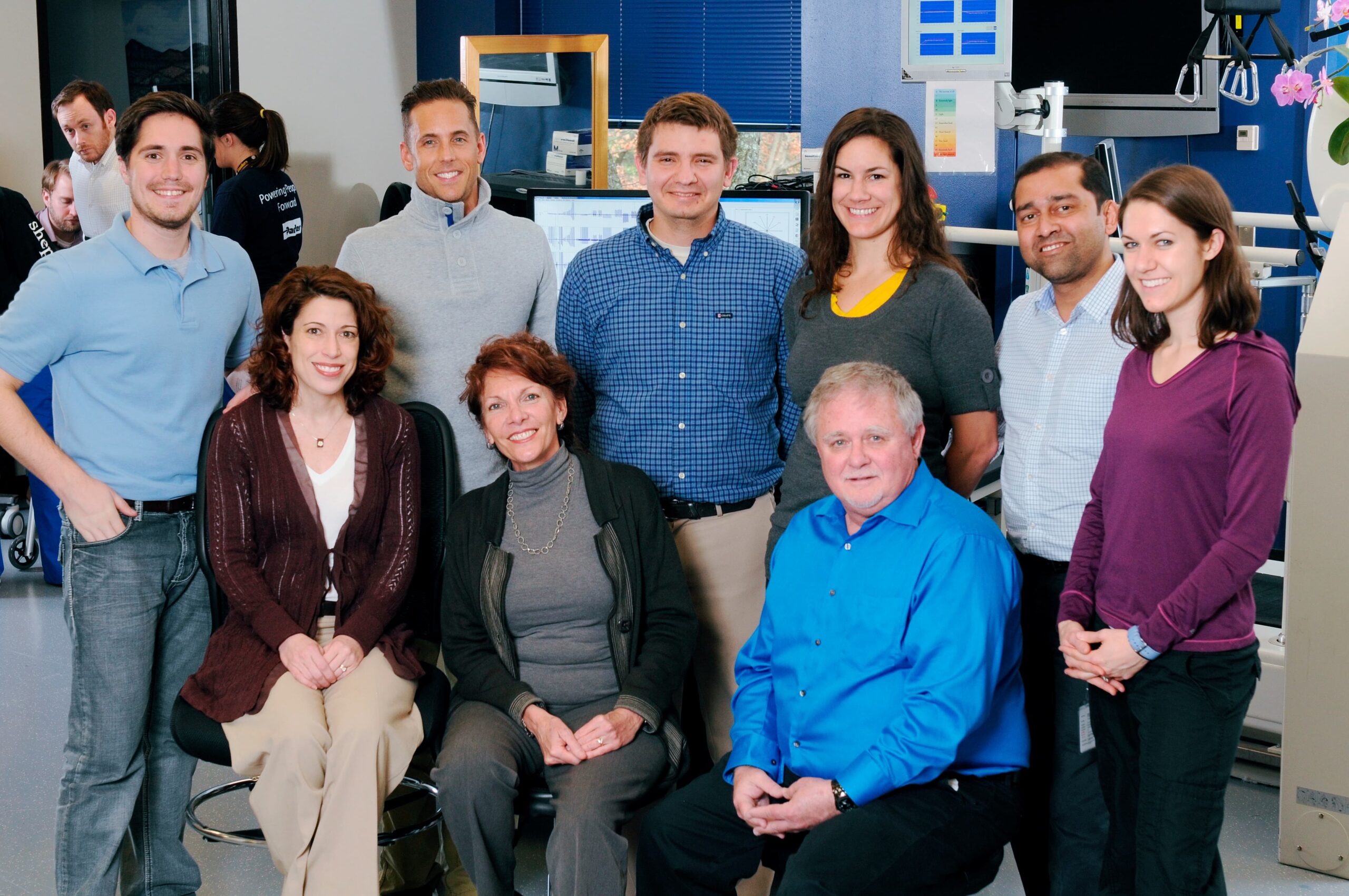 A group of nine people, five standing and four seated, are posing for a photo in a brightly lit office or workspace. They are smiling and dressed in business casual attire. Computers and office equipment are visible in the background.