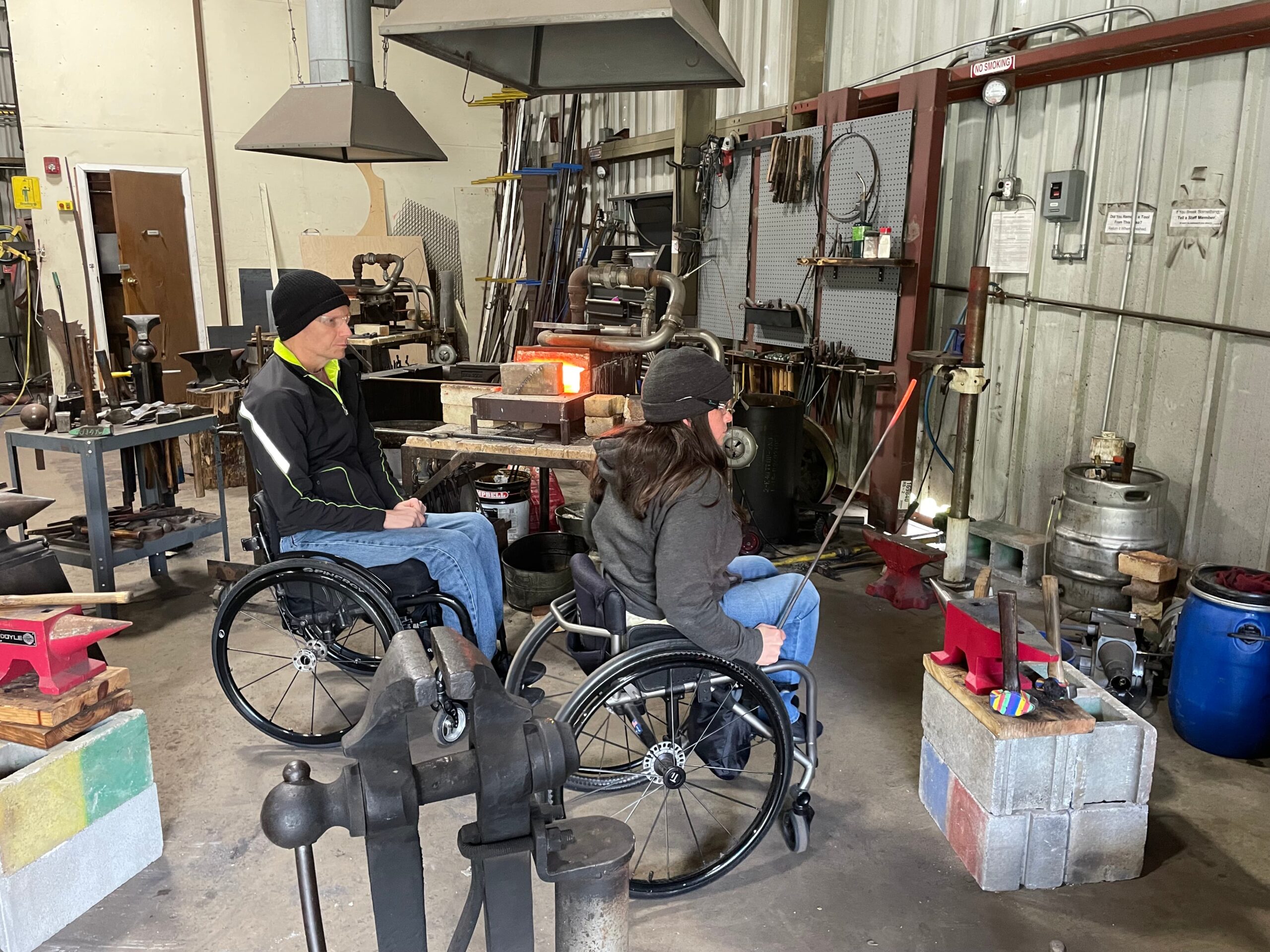 Two people in wheelchairs are observing a blacksmith workshop. They are facing a furnace, with various tools and equipment around them, including anvils and metal rods. The workshop has a rustic and industrial feel.