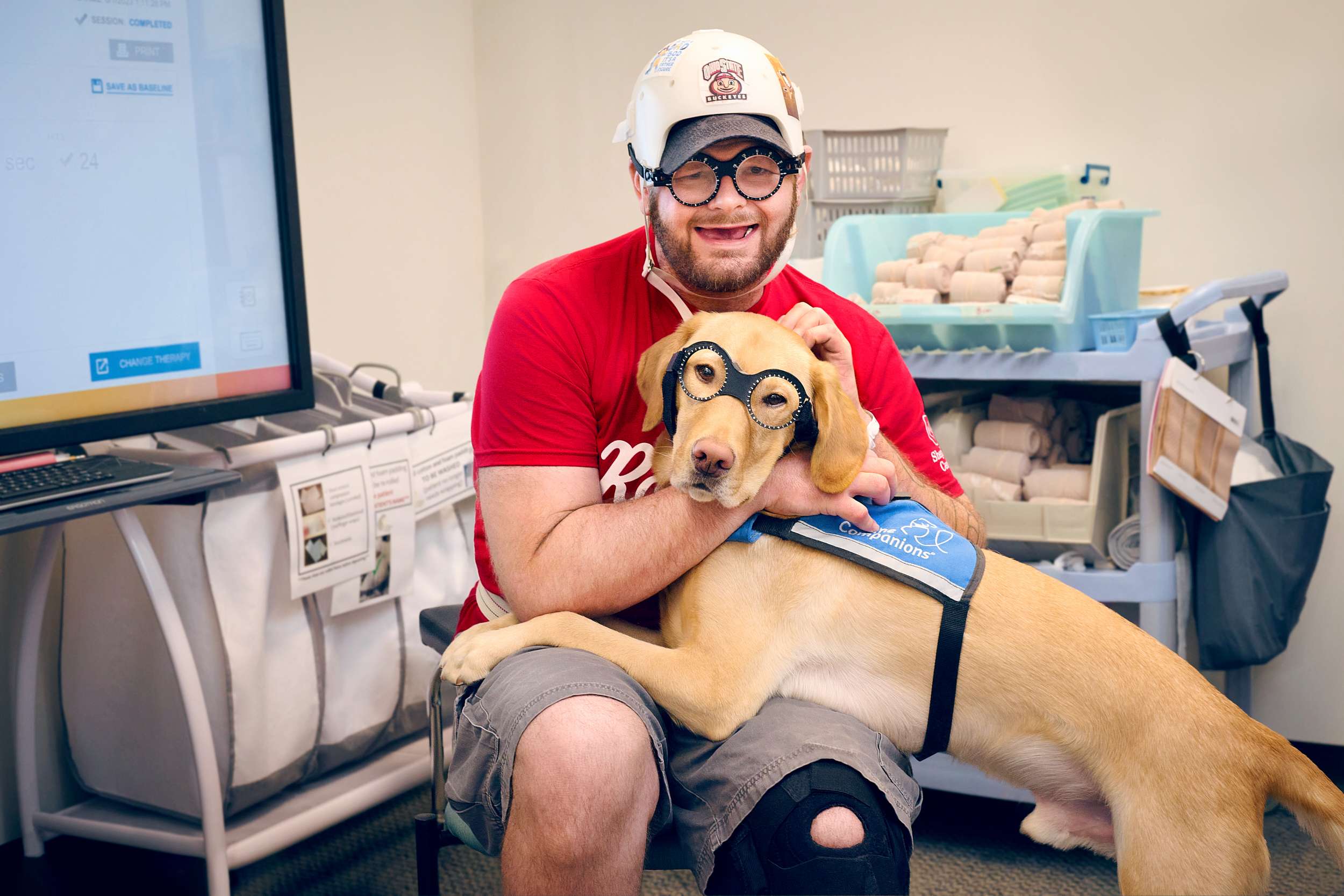 A patient wearing a red shirt, shorts, and protective headgear, smiles and embraces a yellow Labrador retriever wearing a service dog harness. Both are seated in a room with medical supplies and a computer screen in the background.