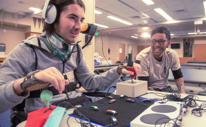 Smiling adolescent boy in a wheelchair and neck brace wearing Microsoft headphones and using the Xbox Adaptive Controller to play video games with a friend.