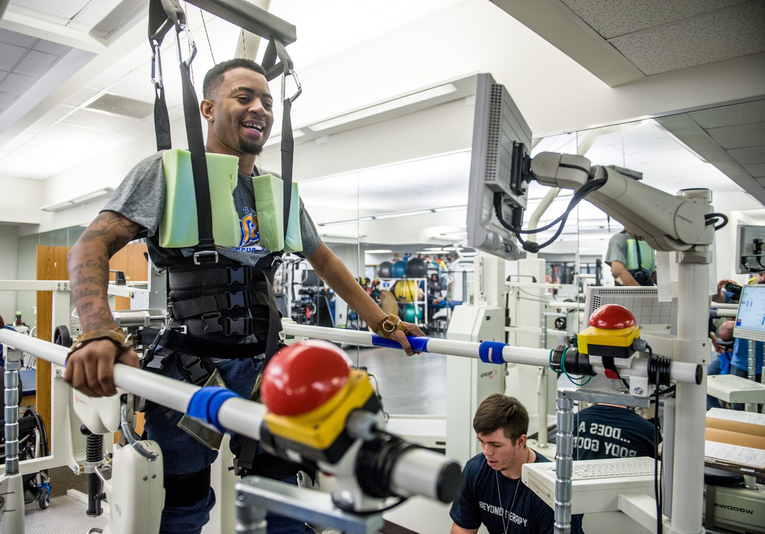 A man wearing a harness and support system is on a treadmill in a rehabilitation center, smiling as he looks at a screen in front of him. A physical therapist kneels nearby, adjusting equipment controls.