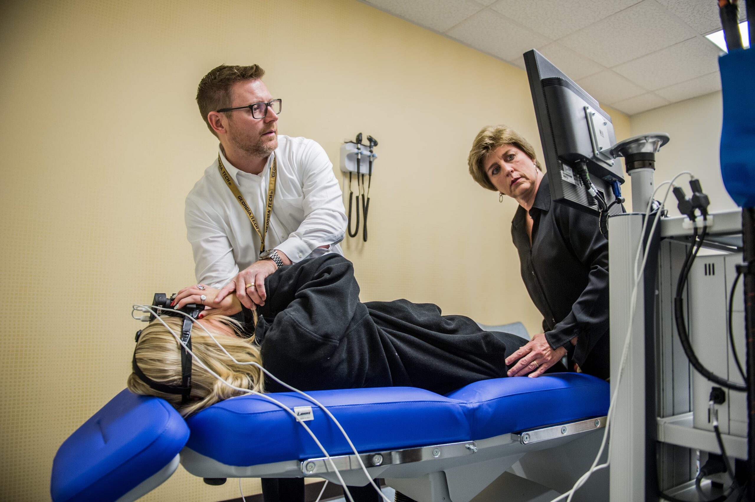 A woman lies on her side on a blue padded examination table with medical sensors attached to her head. A man in a white shirt adjusts the sensors while another woman stands nearby, closely observing a monitor.