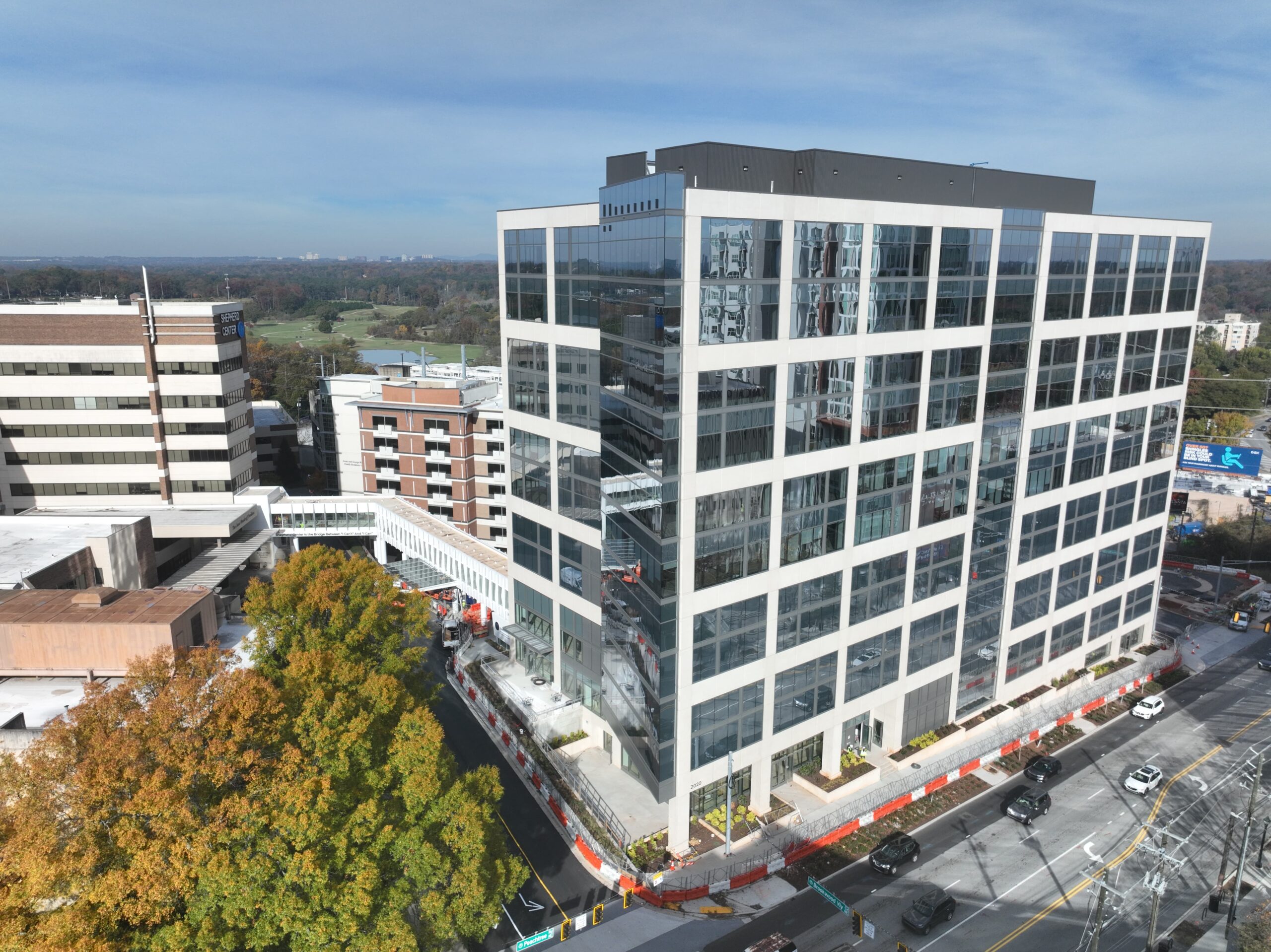 A modern, multi-story office building with large glass windows stands at a street corner. People walk on the sidewalks, and cars are on the road. The sky is clear and blue, and nearby trees add greenery to the urban scene.