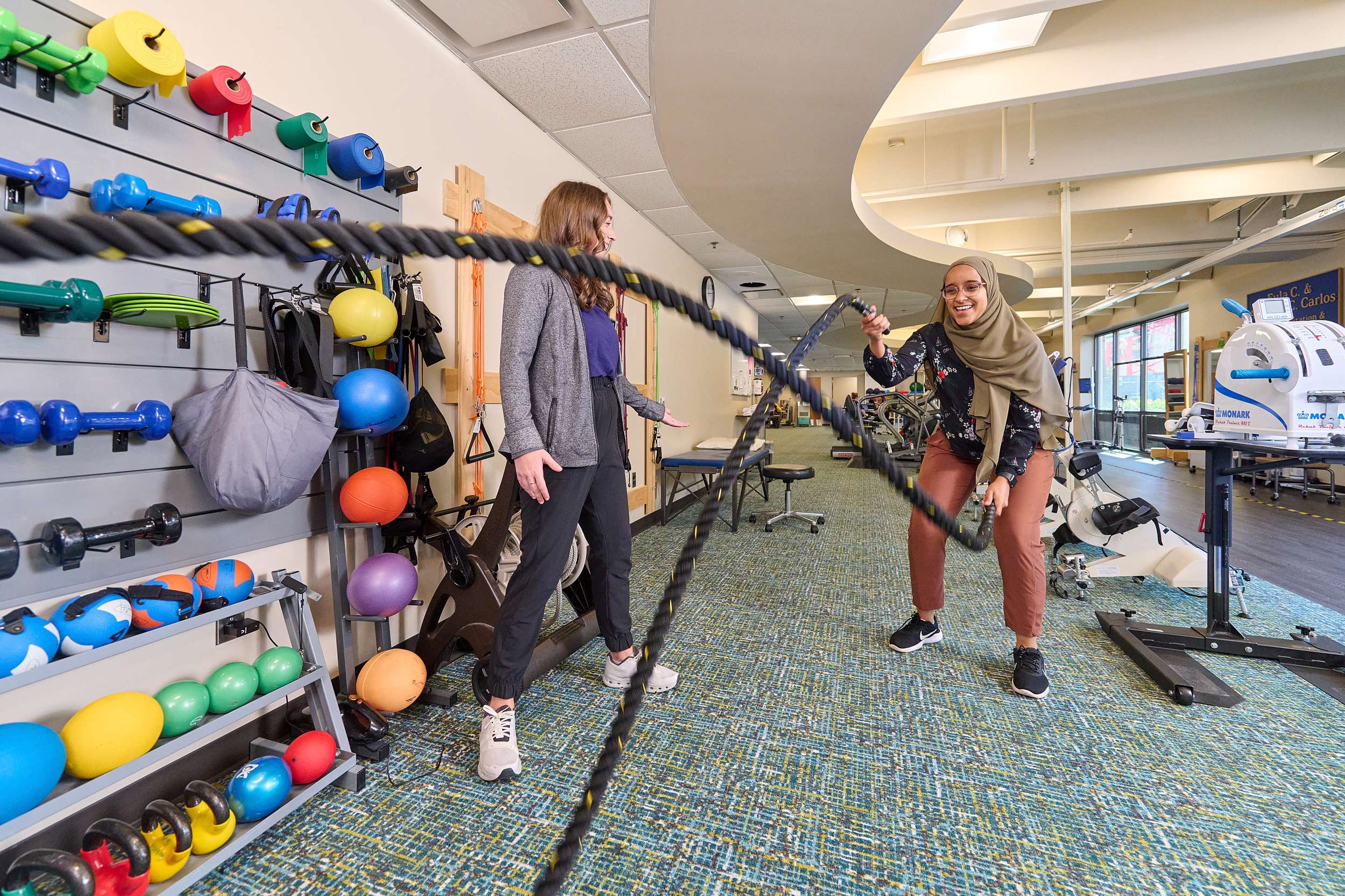 Two women are engaged in a workout using battle ropes in a gym. One is holding and swinging the ropes, while the other stands beside her, observing and smiling. The gym has various fitness equipment such as dumbbells, exercise balls, and resistance bands on the wall.