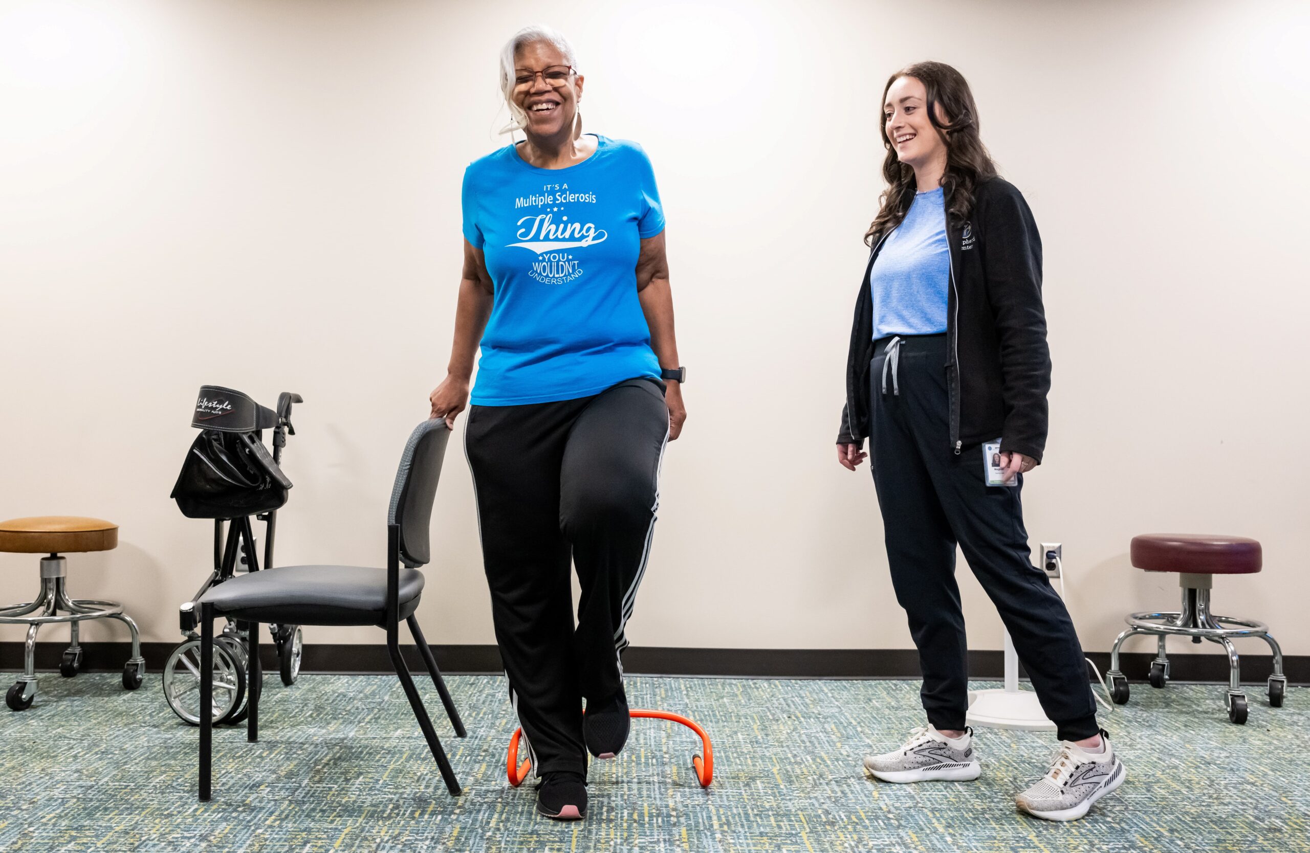 An older woman smiles as she performs a leg exercise using a chair in a room with a walker and stools. She wears a blue shirt and black pants. A young female instructor, also smiling, stands nearby, wearing a light blue shirt and black jacket.