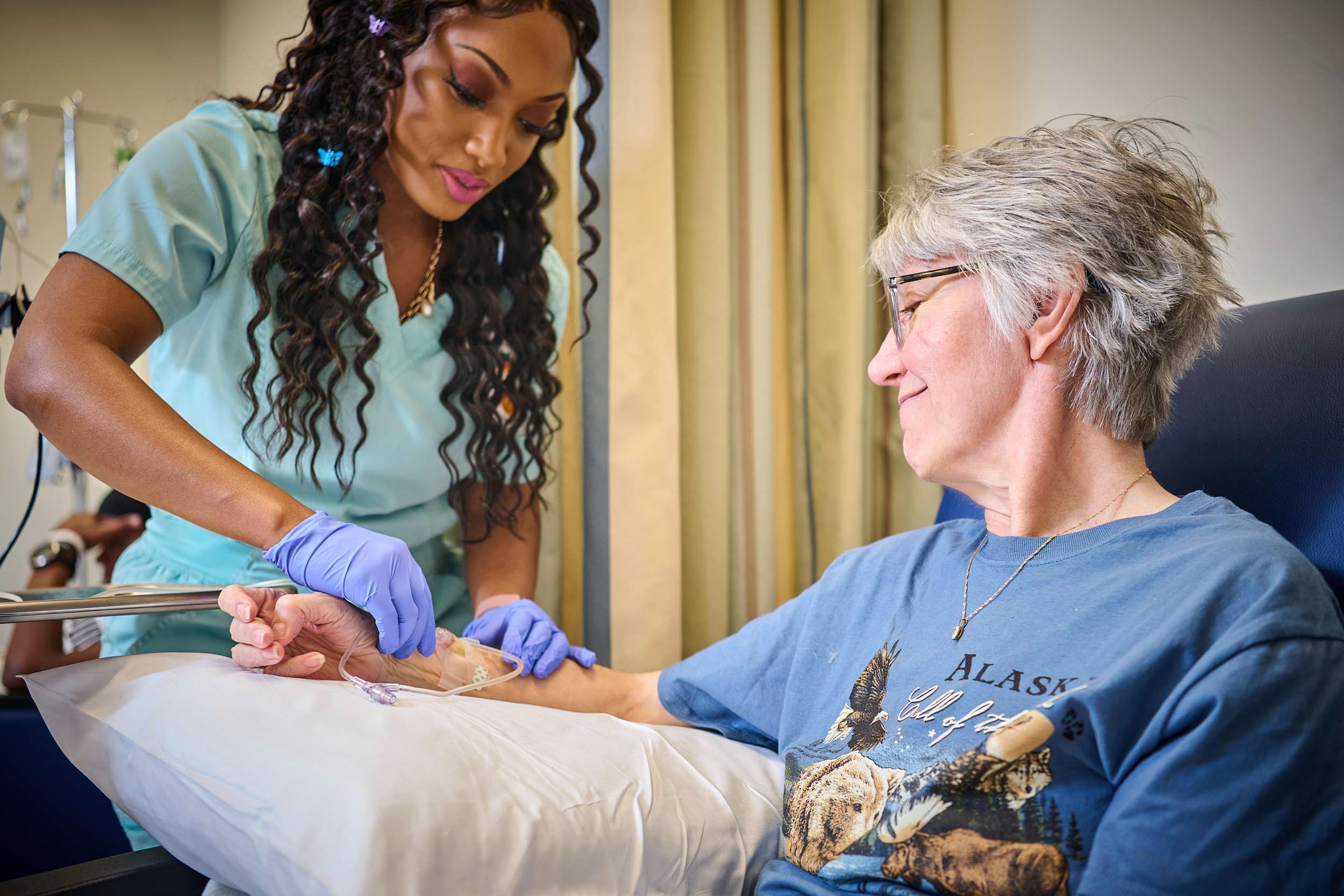 A healthcare professional in light blue scrubs and purple gloves administers an IV to an older woman sitting in a chair. The woman, wearing glasses and a T-shirt with an Alaska design, smiles and looks at the healthcare professional.