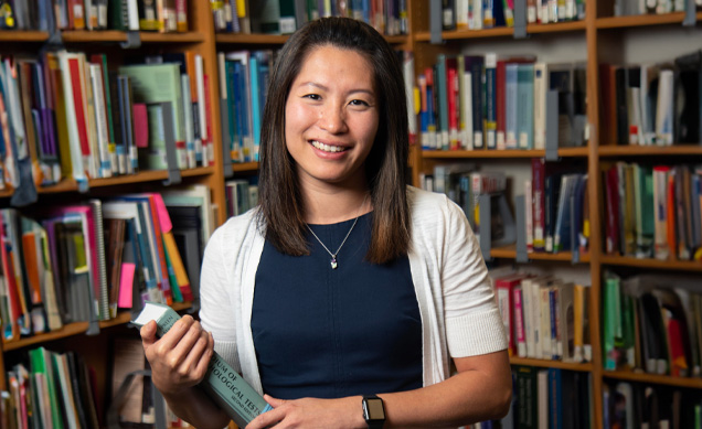 A woman with shoulder-length hair stands in a library, smiling. She is wearing a white cardigan over a dark top and holds a book. Shelves filled with books are visible in the background.