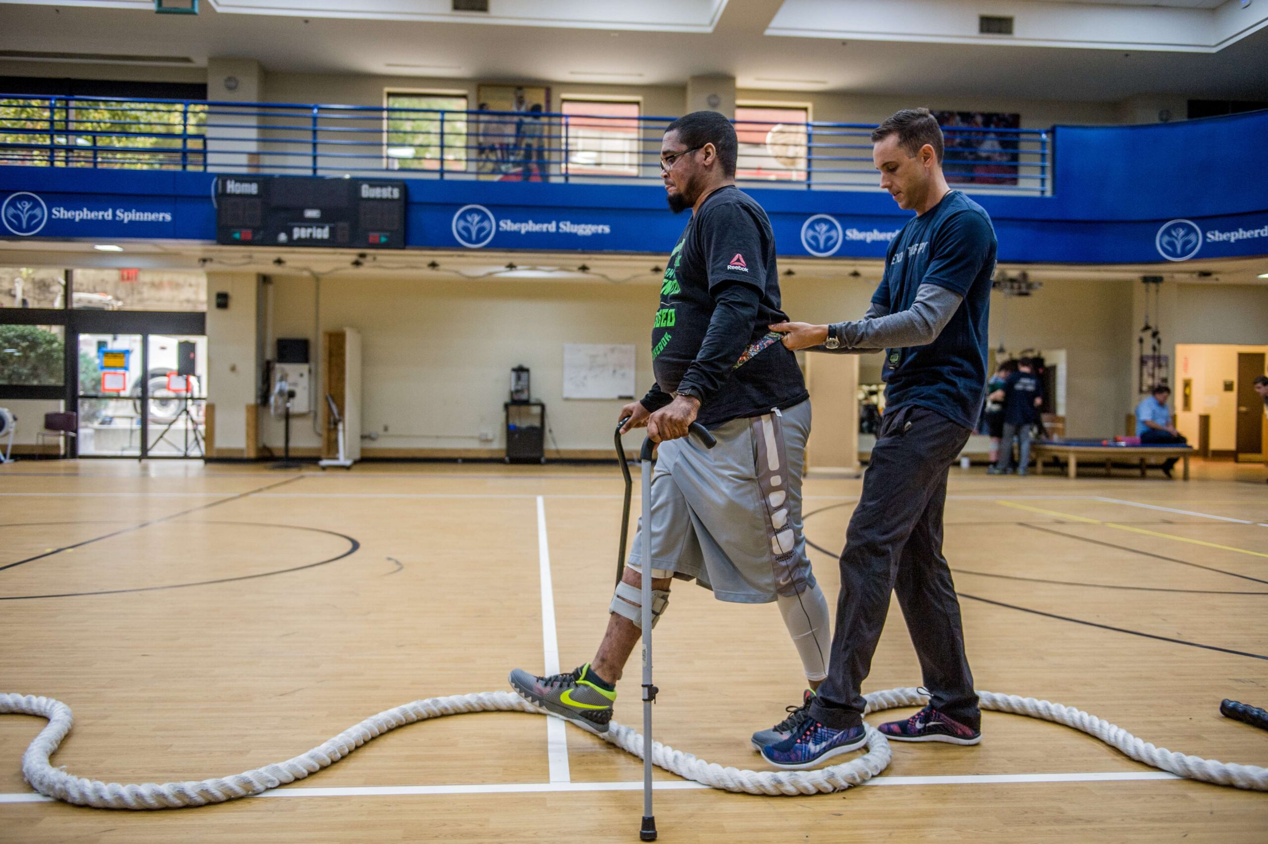 A man is walking across a gym floor with the aid of a walking cane, while a physical therapist closely supports him from behind. The gym has a blue railing on the upper level and various equipment visible in the background.