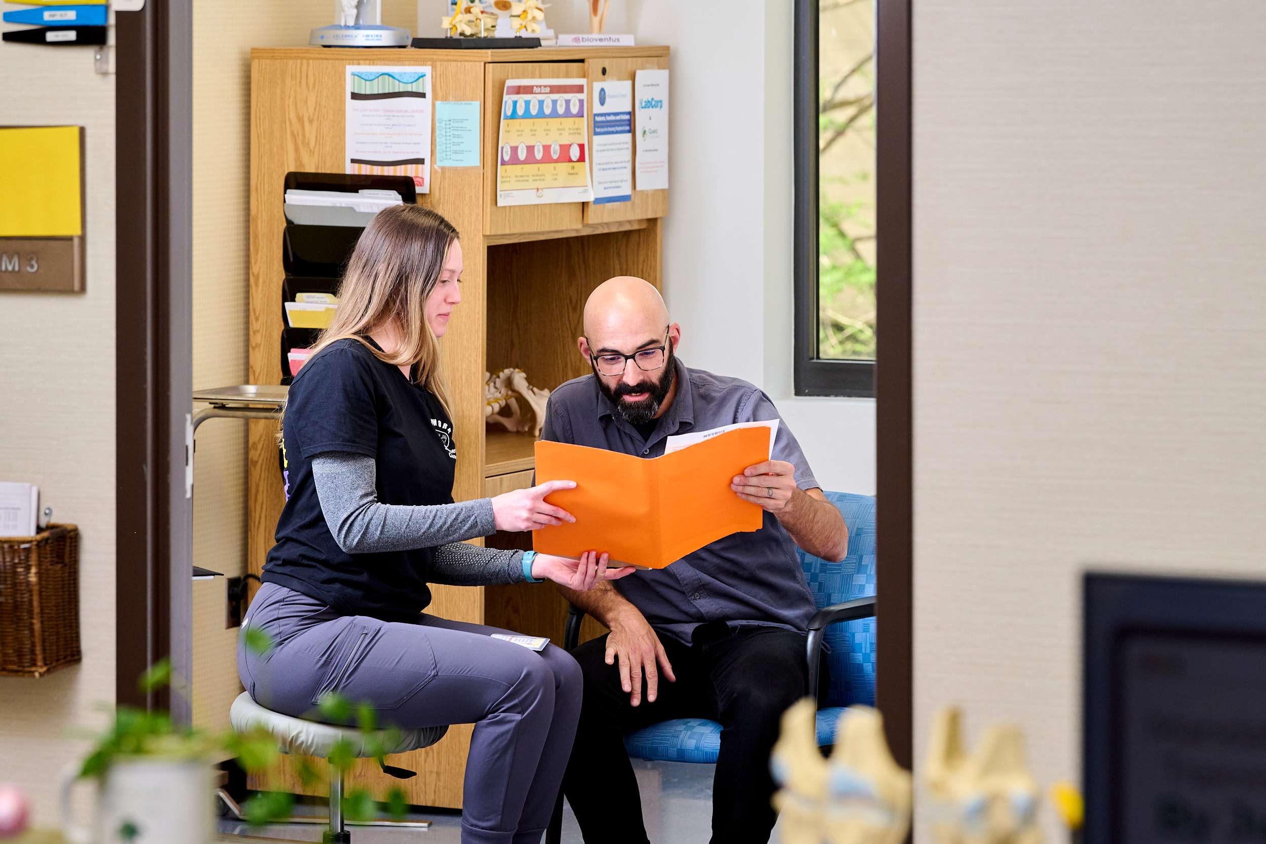 A woman and a man sitting in an office, reviewing documents in an orange folder. The woman is dressed in casual attire, and the man is wearing a dress shirt. They appear engaged in discussion. Shelves and informational posters are visible in the background.