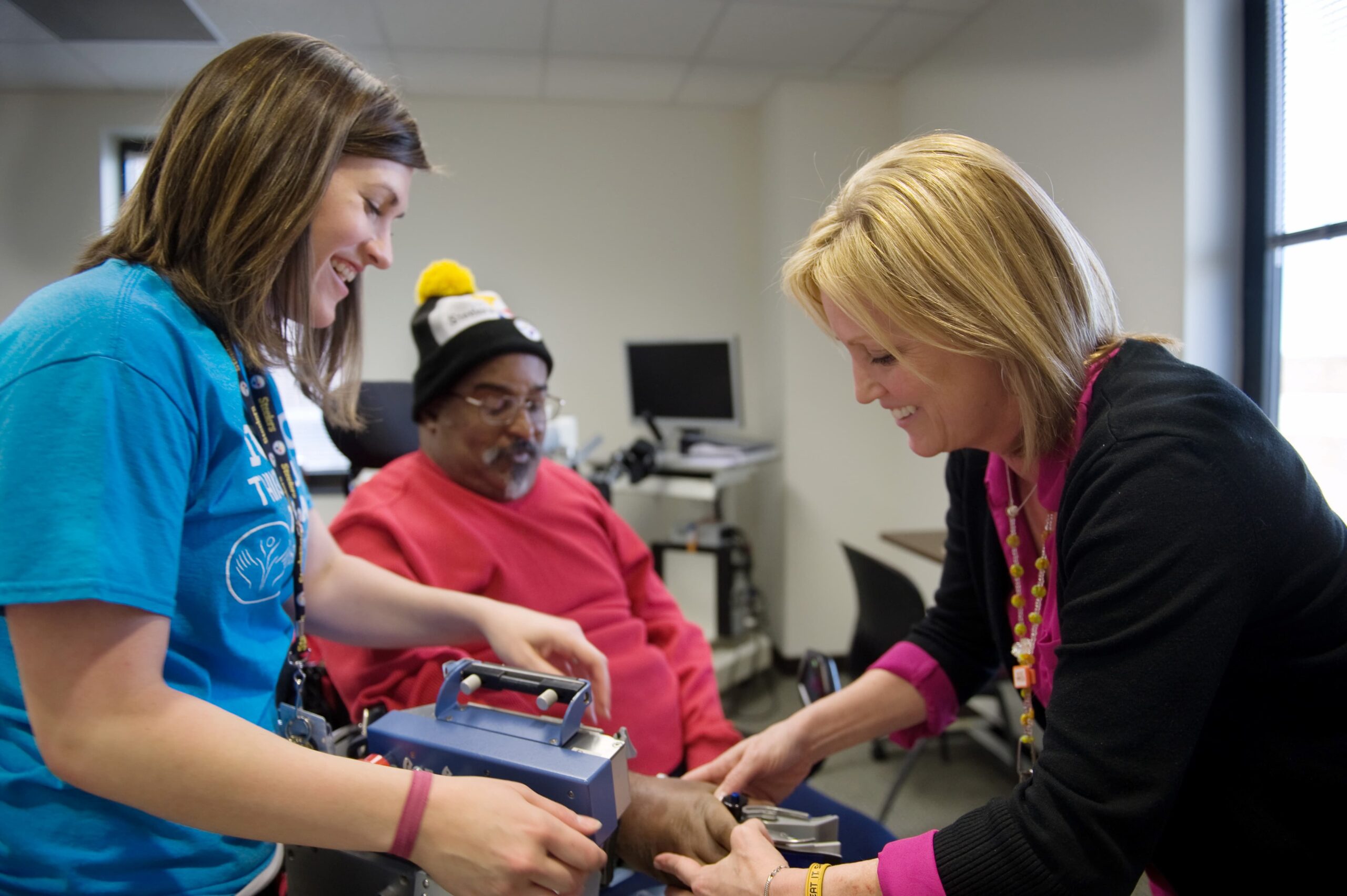 Two healthcare professionals examine the arm of an older man sitting in a chair in a medical office. The man wears a red sweater and a winter hat.