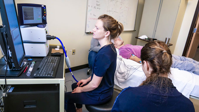 Three medical professionals are conducting a sleep study. One person is monitoring a computer screen displaying data, while a second person observes. A patient lies on a bed in the background, connected to various sensors. A whiteboard with notes is in the background.