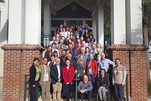 A large group of people posing on the steps of a building with columns, some sitting and others standing. Most are wearing business-casual attire, and many are smiling at the camera. The building has a covered entrance made of brick and white columns.