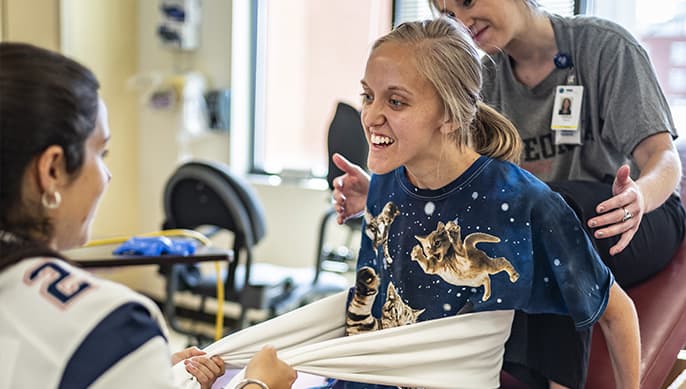 A young woman with a bright, joyful expression is assisted by two therapists as she participates in a physical therapy session. The woman is holding onto a white resistance band, and one therapist is supporting her back while the other is in front of her.