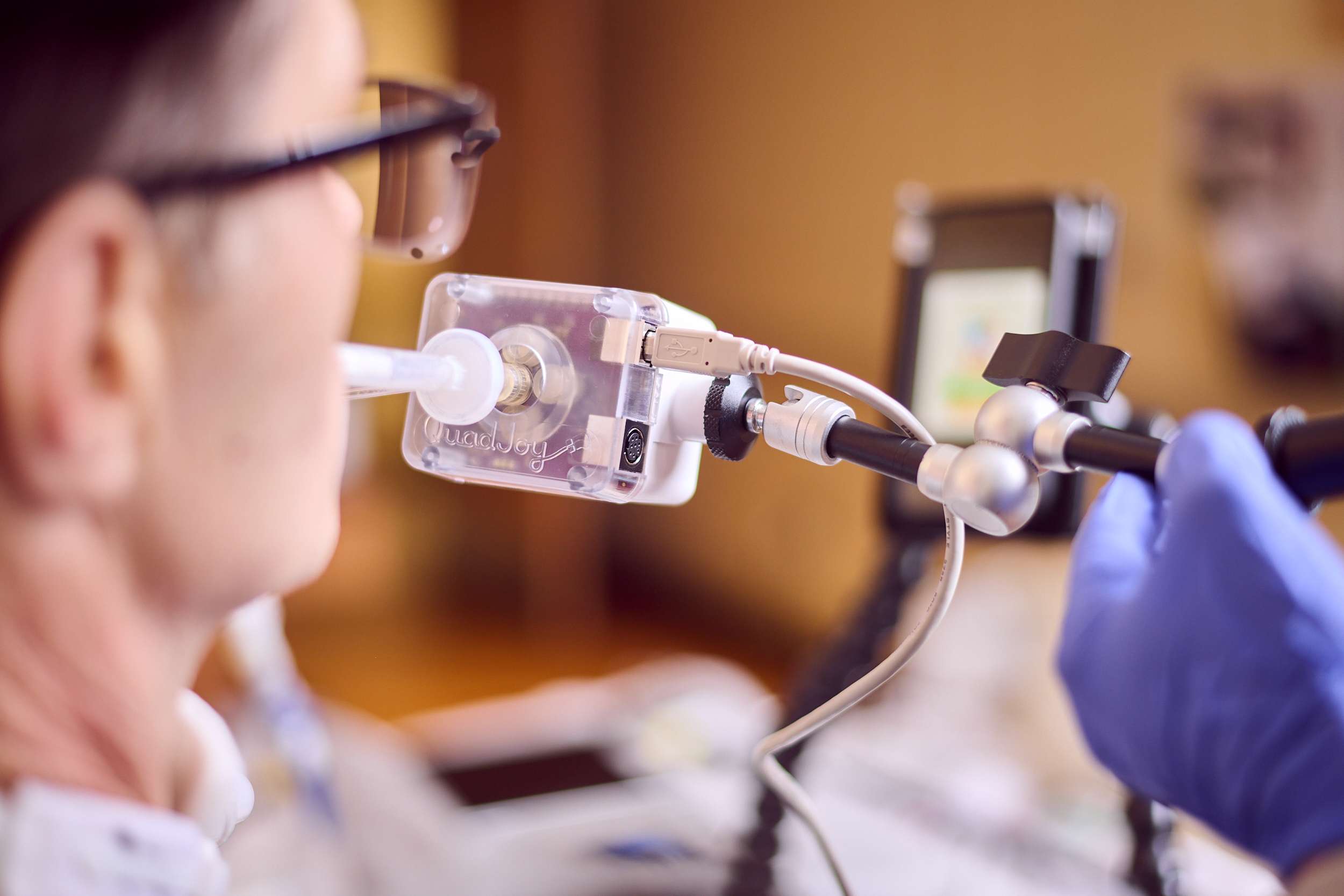 A close-up shot of a person using assistive technology: a breath-controlled device with a mouthpiece attached to a stand, connected to a mobile phone. A person wearing glasses is positioned to use the device, and a gloved hand adjusts the setup.