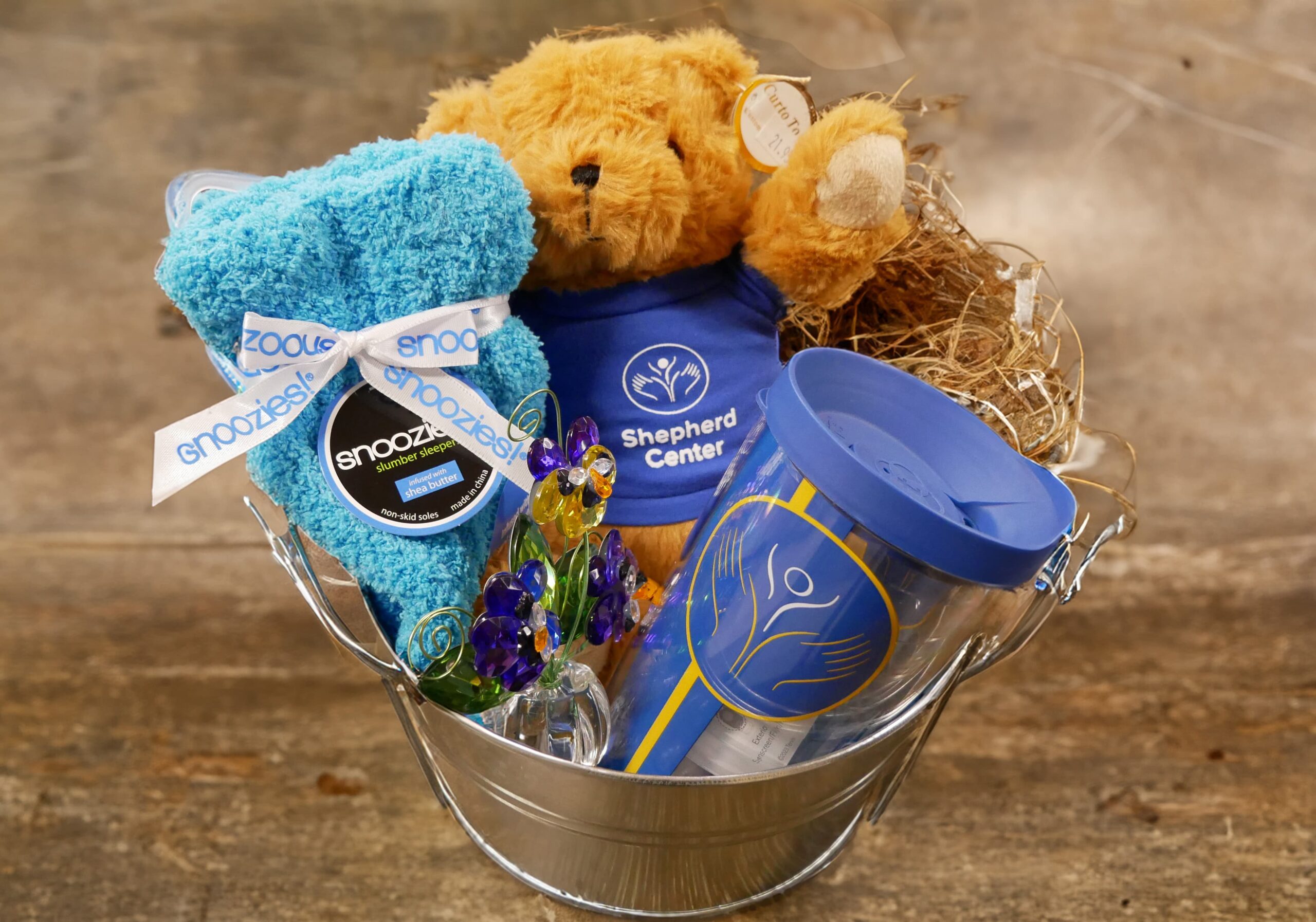 A silver basket contains a teddy bear wearing a blue shirt, a blue tumbler, a plush rolled towel with a ribbon, and decorative flowers. The basket rests on a wooden surface.