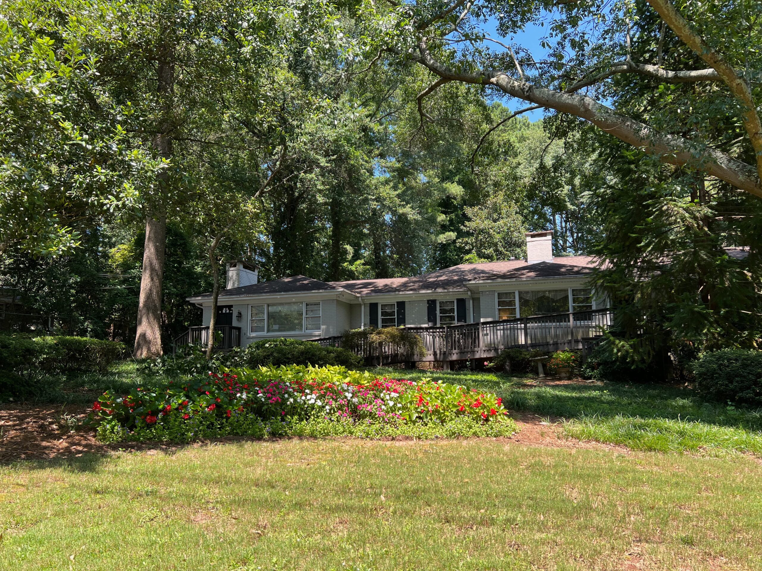 A charming single-story house surrounded by lush green trees on a sunny day. The front yard features a colorful flower bed with red and white blossoms, adding vibrant contrast to the natural setting. A wooden deck is visible along the front of the house.