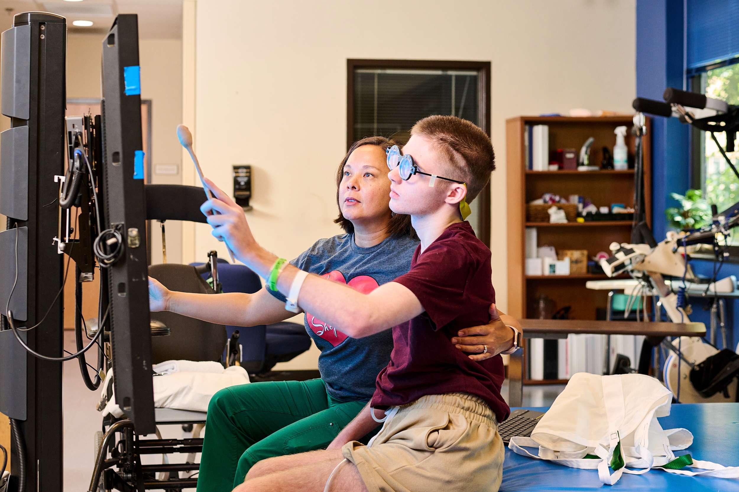 An occupational therapist sits next to a young man wearing Prism glasses as he uses a touch screen.