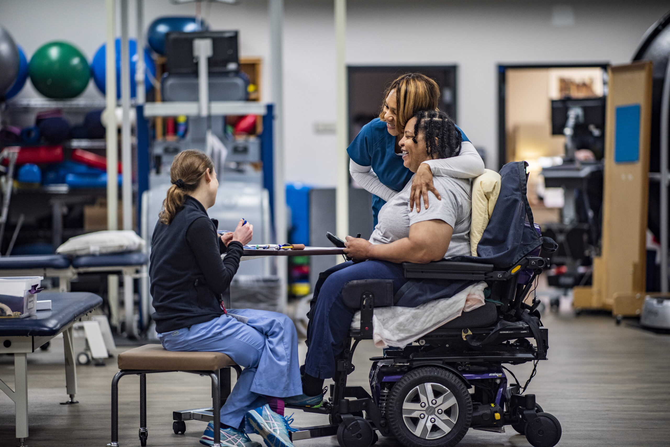 A therapist in scrubs, seated, talks with a patient in a wheelchair, while another staff member standing nearby hugs them warmly. The setting is a physical therapy facility with exercise equipment in the background.