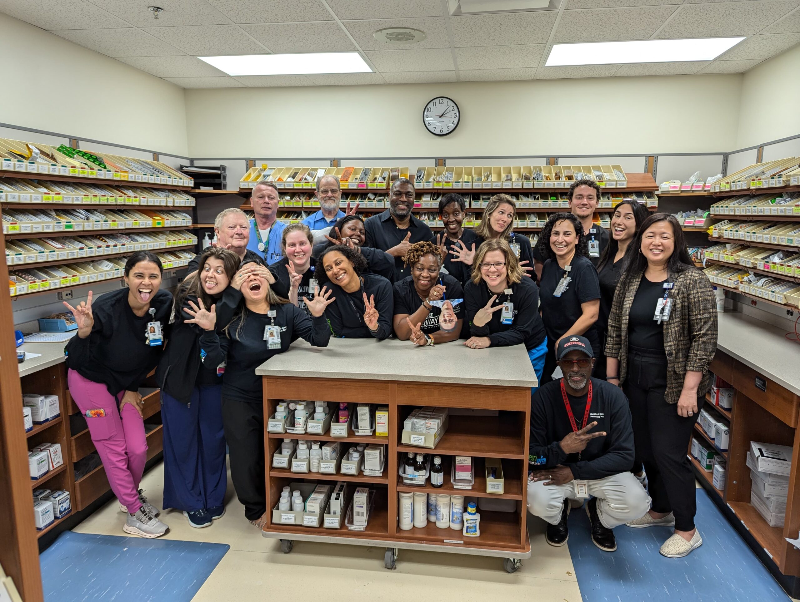 A diverse group of people wearing uniforms pose together in a pharmacy setting. They are gathered around a counter, with rows of medication shelves in the background. Some are making playful gestures and everyone is smiling.