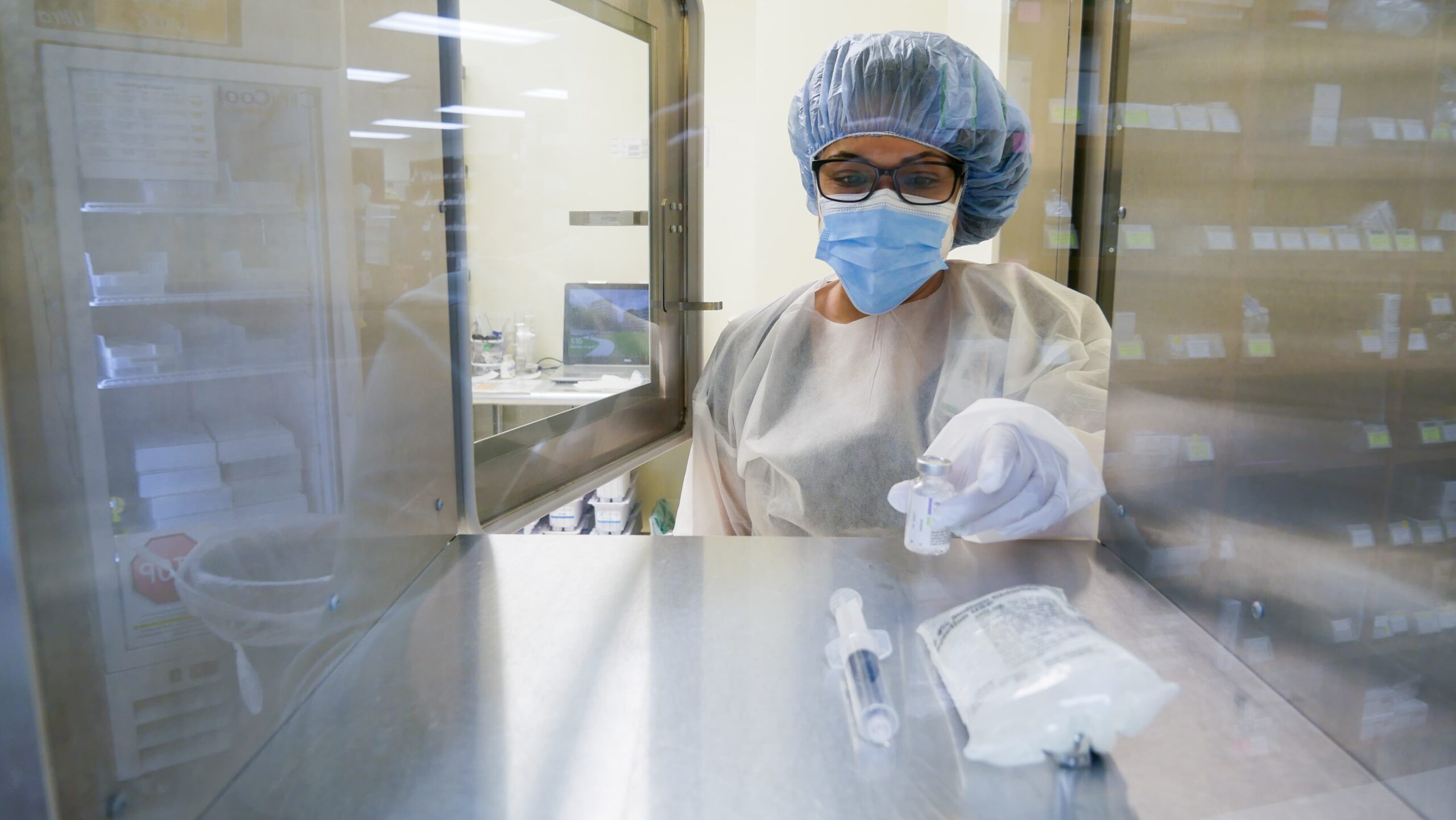 A healthcare worker in protective gear prepares a syringe and medicine vial in a sterile environment. They are wearing a face mask, hair net, and gloves, focusing intently on their task through a safety barrier.