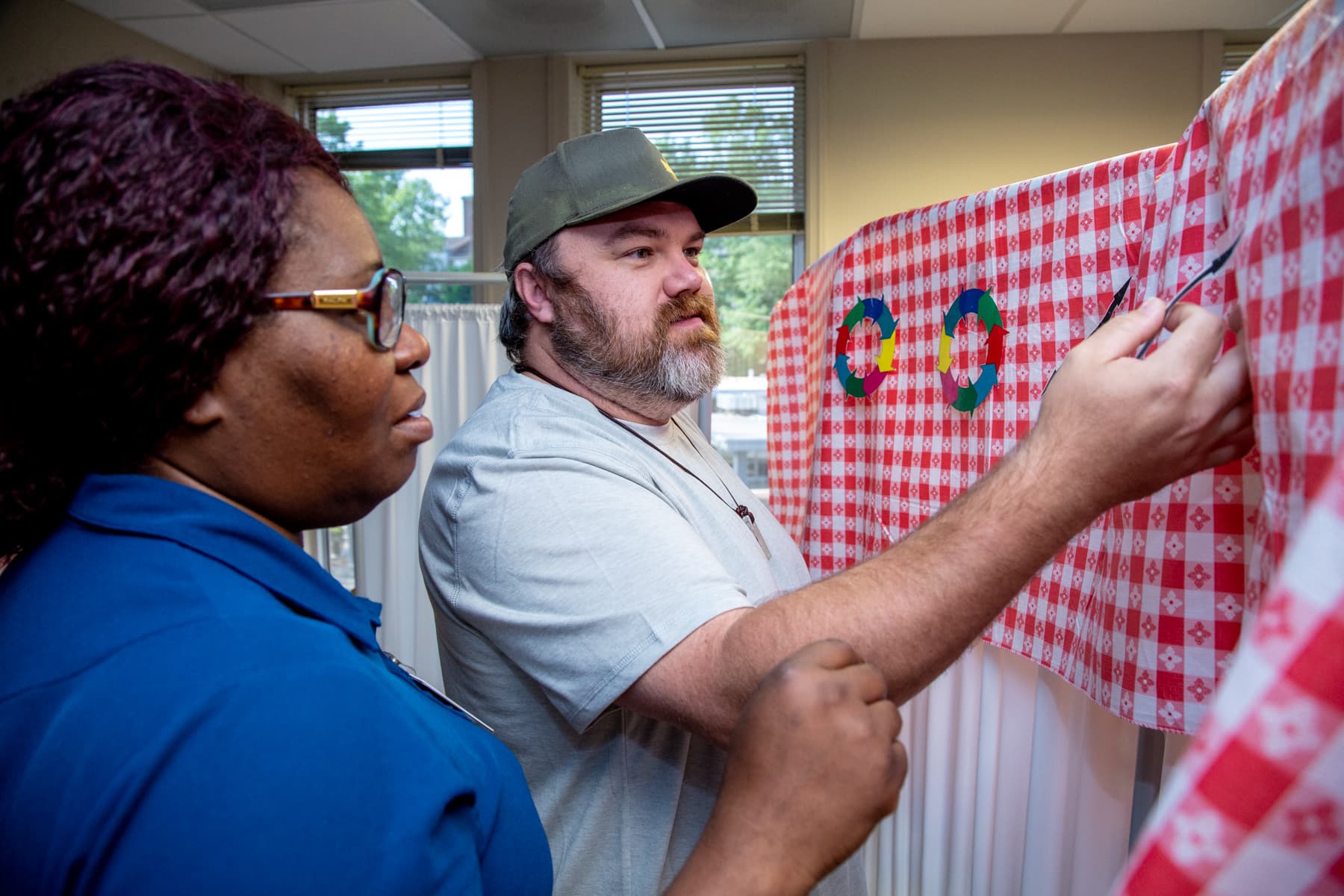 Two people interacting with a checkered cloth hanging on a wall. The person on the left wears glasses and a blue top. The person on the right has a beard, wears a hat and a light gray top, and is holding a writing instrument while gesturing towards the cloth.