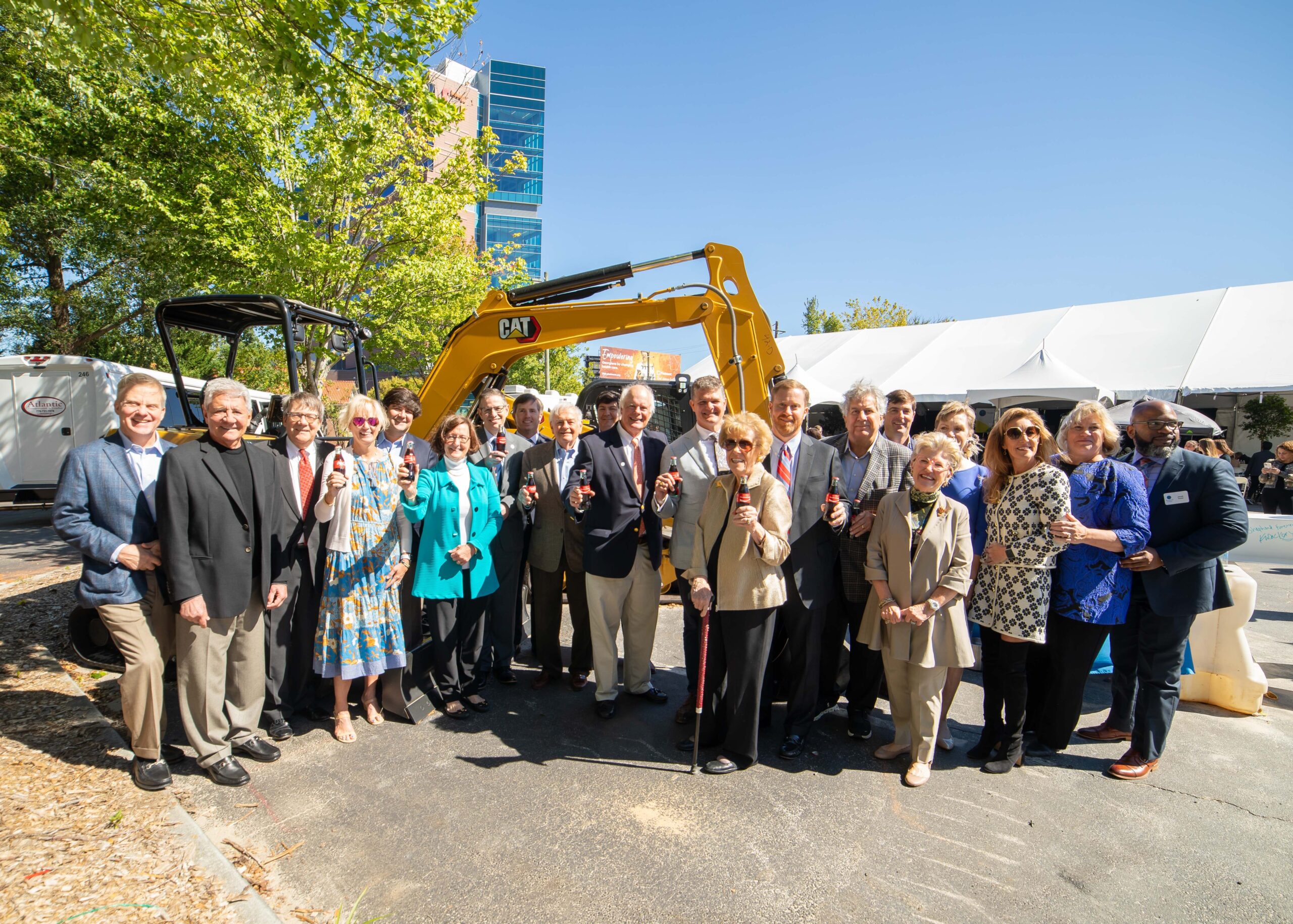 A group of people stand in front of a yellow excavator, holding bottles, at an outdoor event. Some wear suits and others wear casual attire. A white tent and green trees are visible in the background under a clear blue sky.